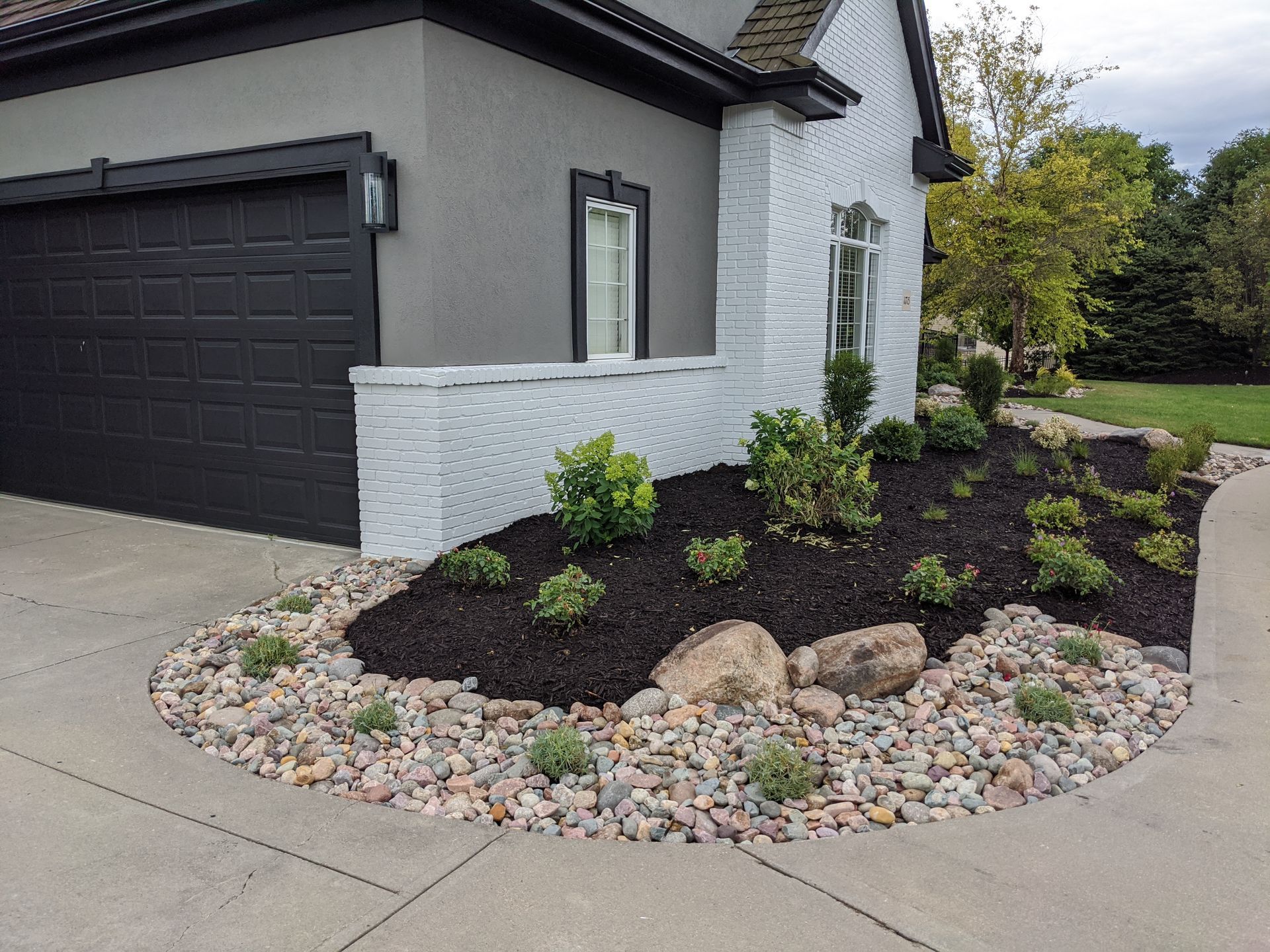 Exterior of a house with a black garage door and a landscaped garden bed with mulch, rocks, and shrubs.