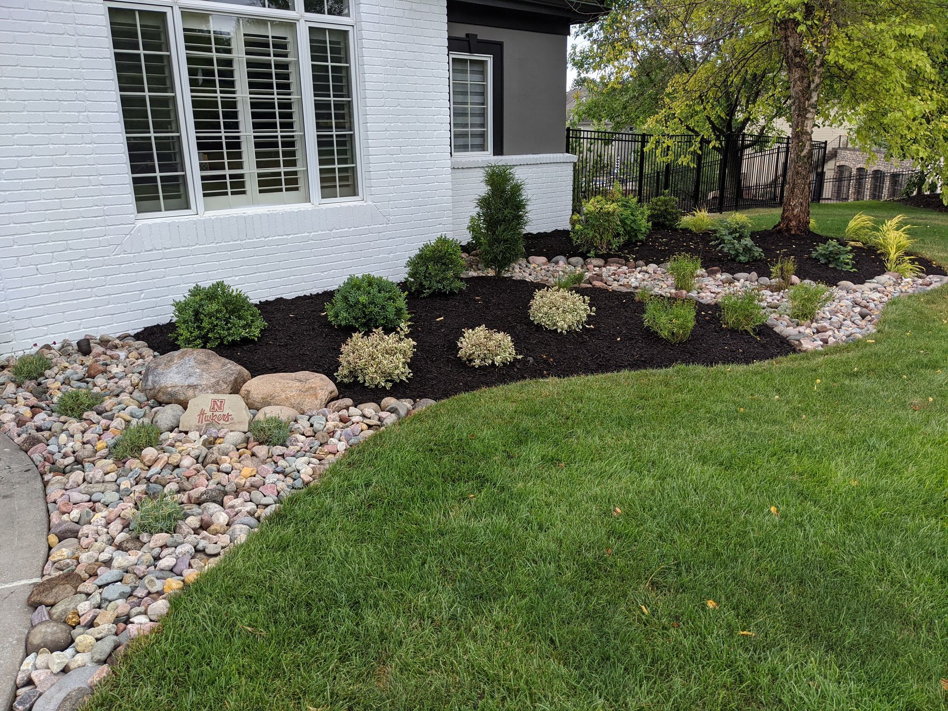 Landscaped yard with grass, mulch, rocks, and bushes next to a white brick house.