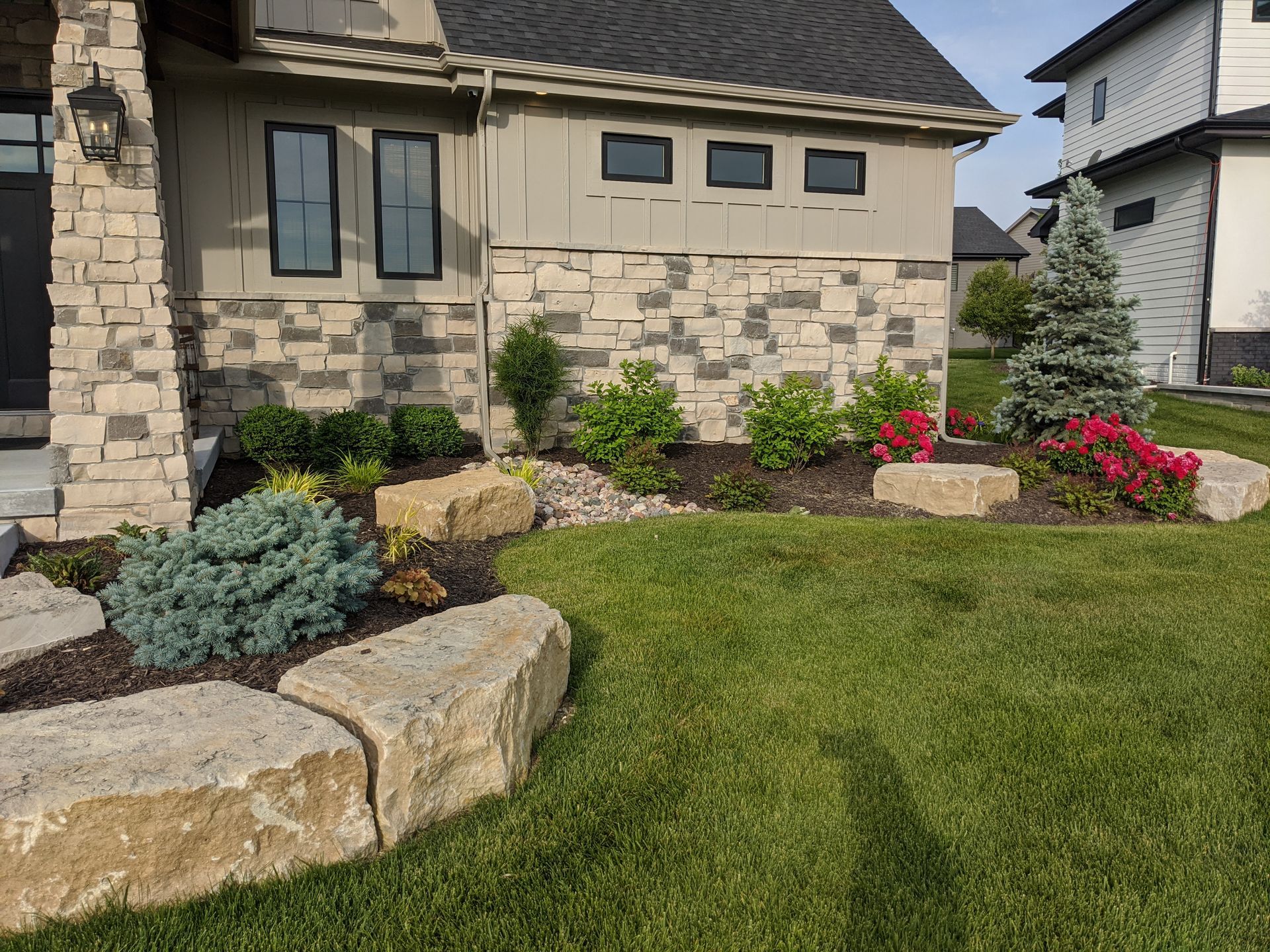 Landscaped front yard with rock edging, various green and red plants, and a gray house.