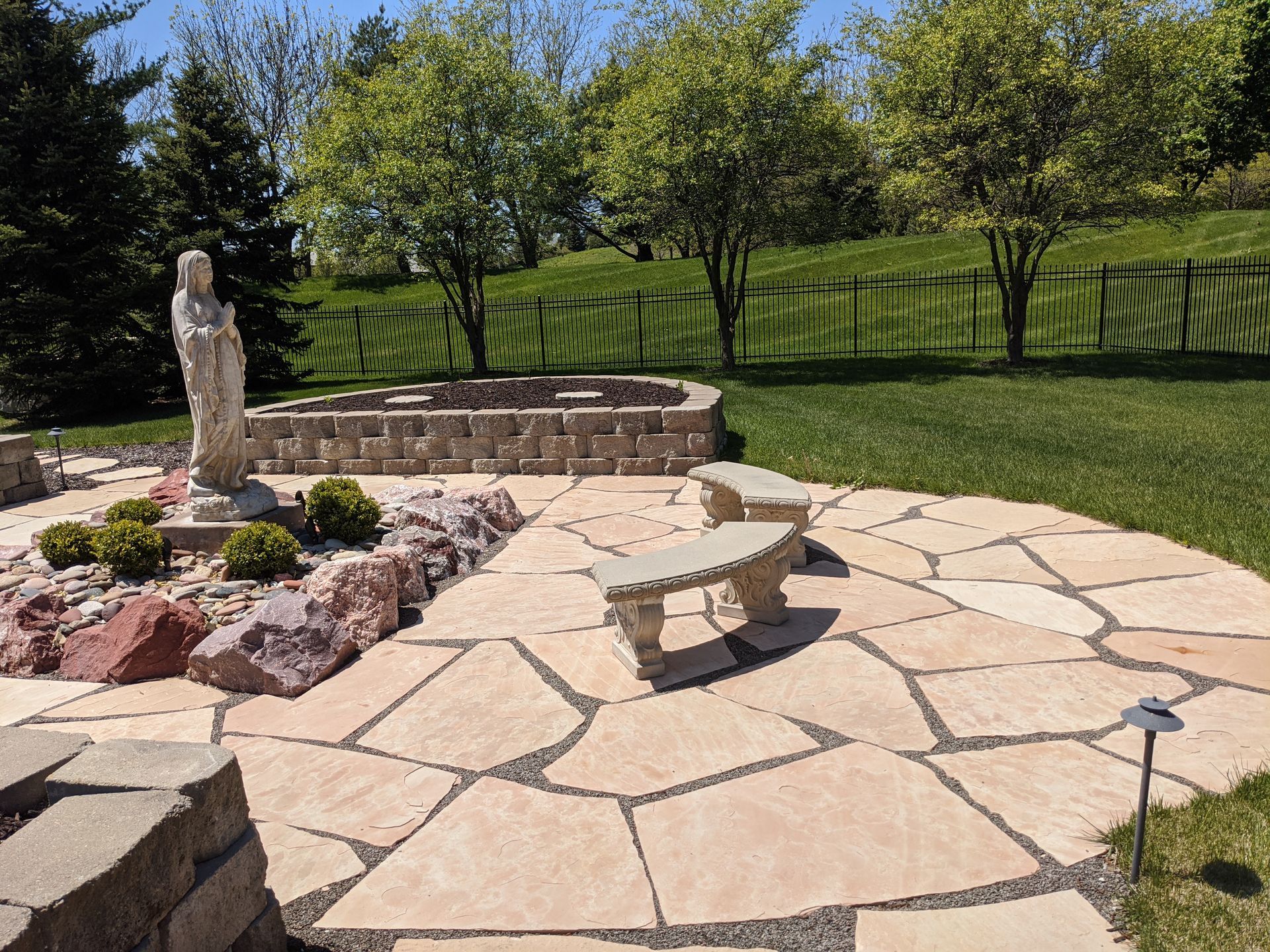 Stone patio with benches, statue, and fire pit in a sunny backyard with trees and green grass.
