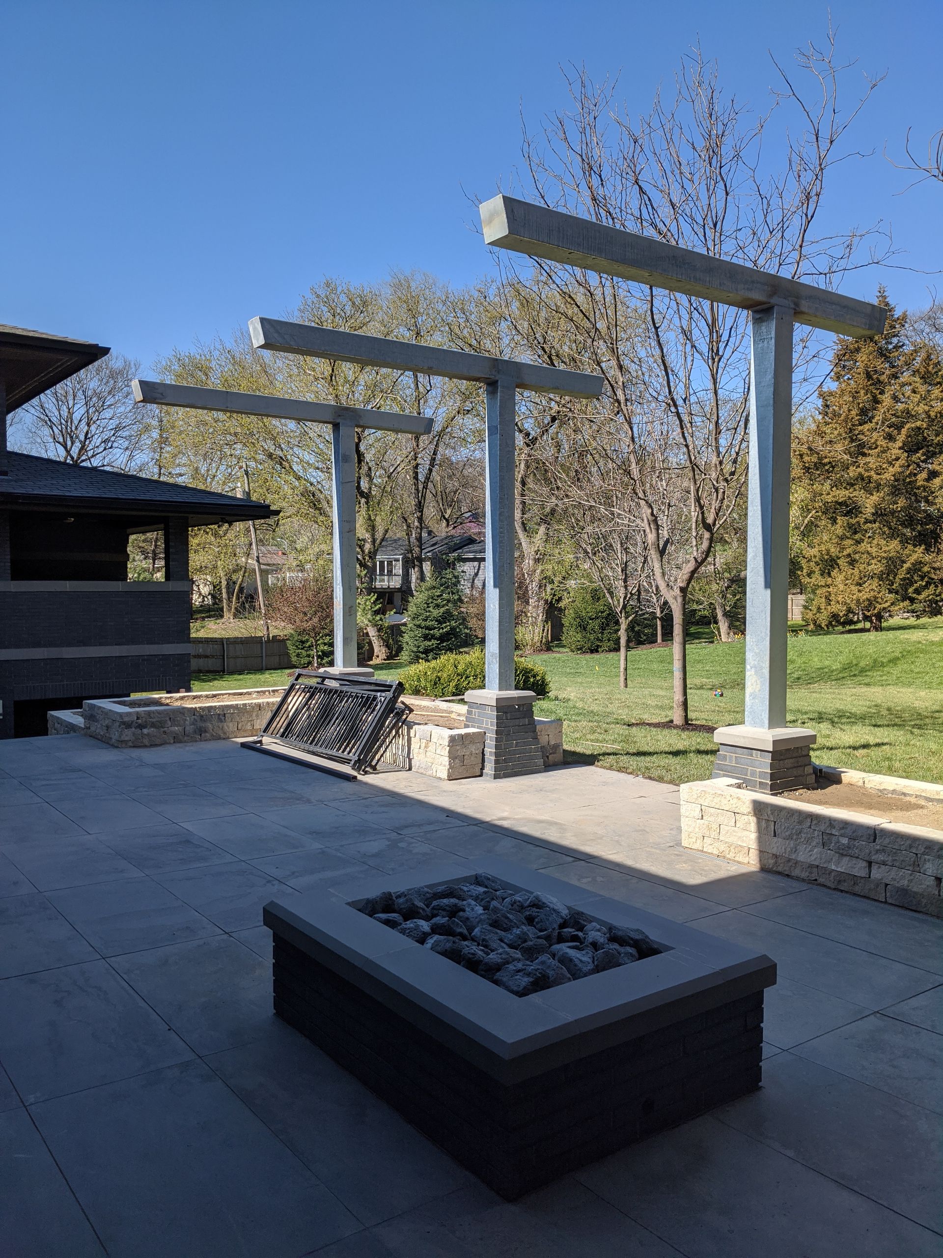 Outdoor patio with fire pit, pergola, and low retaining walls on a sunny day.