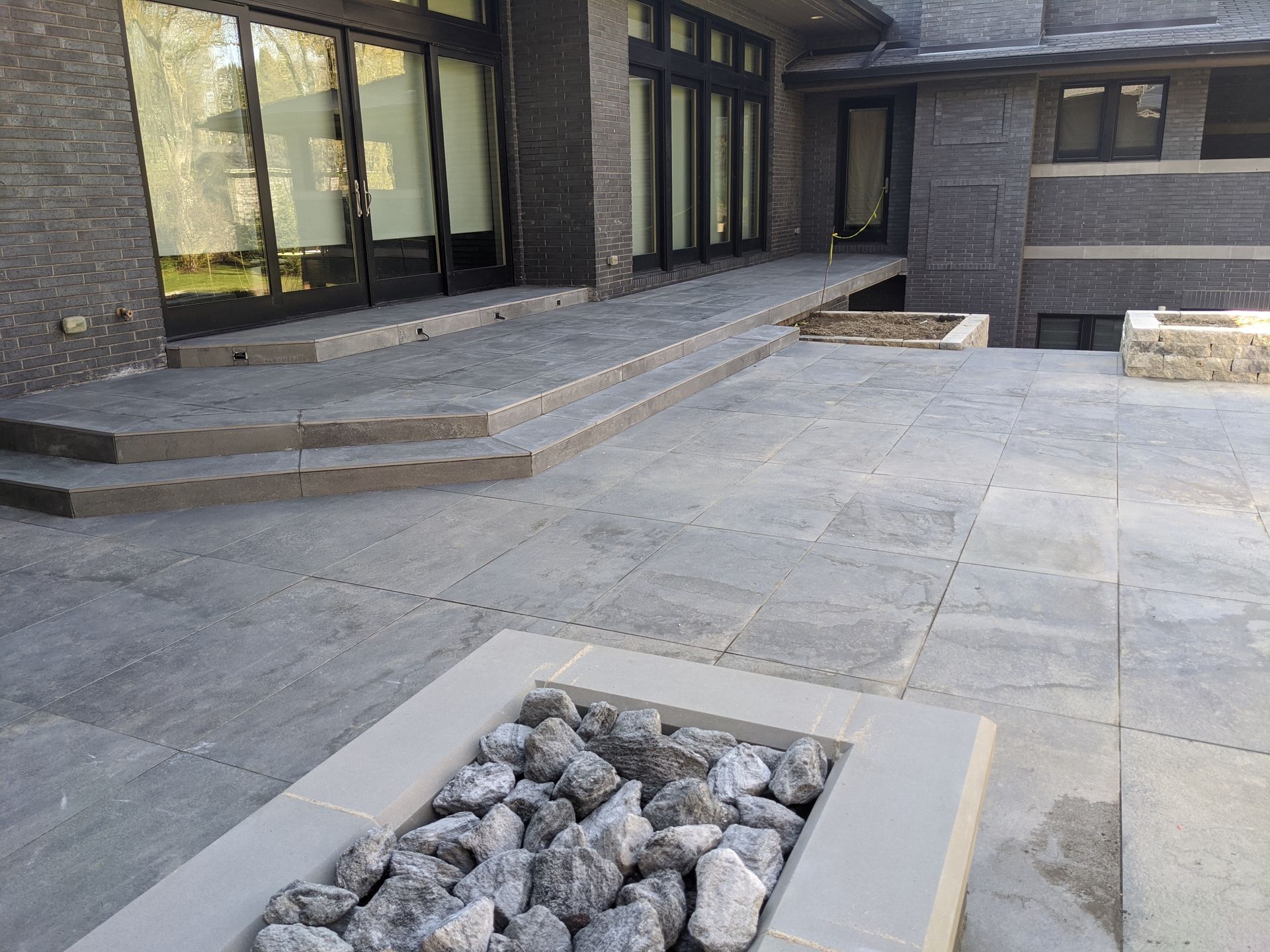Outdoor patio with gray stone flooring, steps, a ramp, and a fire pit filled with rocks. Building with dark brick.