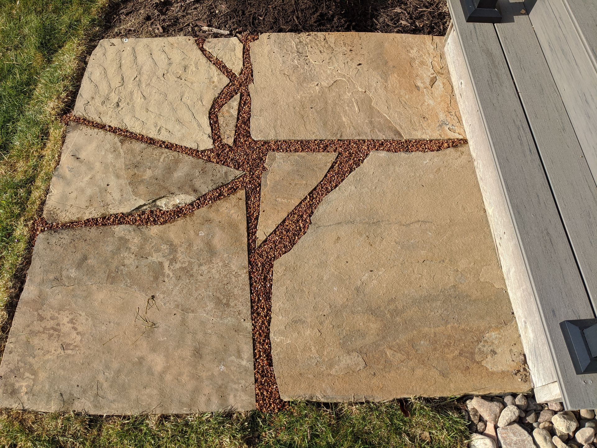 Stone patio with brown gravel-filled seams, next to a deck and green grass.