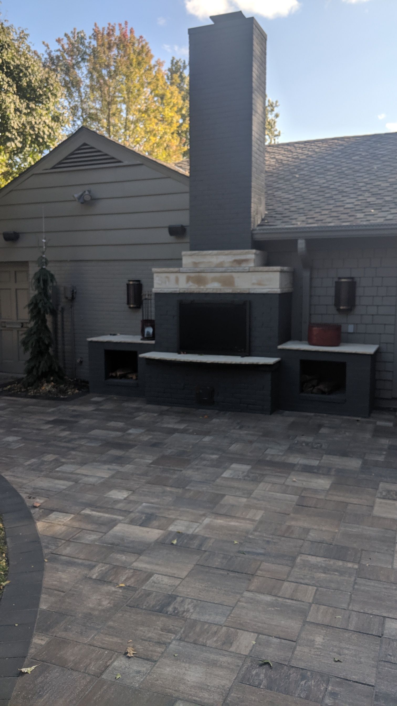Outdoor fireplace with dark gray chimney, brickwork patio, gray house.