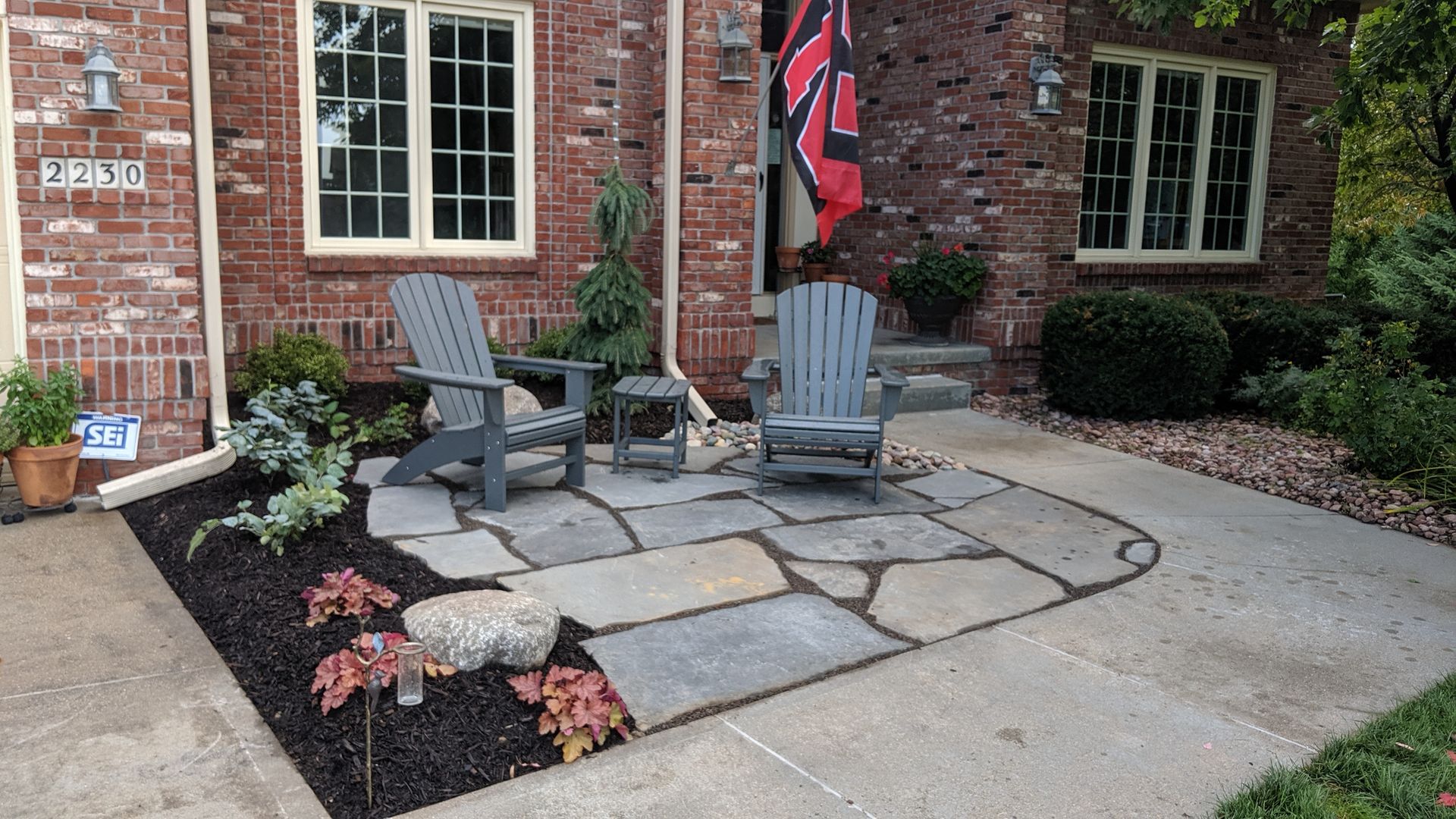 Two gray Adirondack chairs on a stone patio in front of a brick house with landscaping.