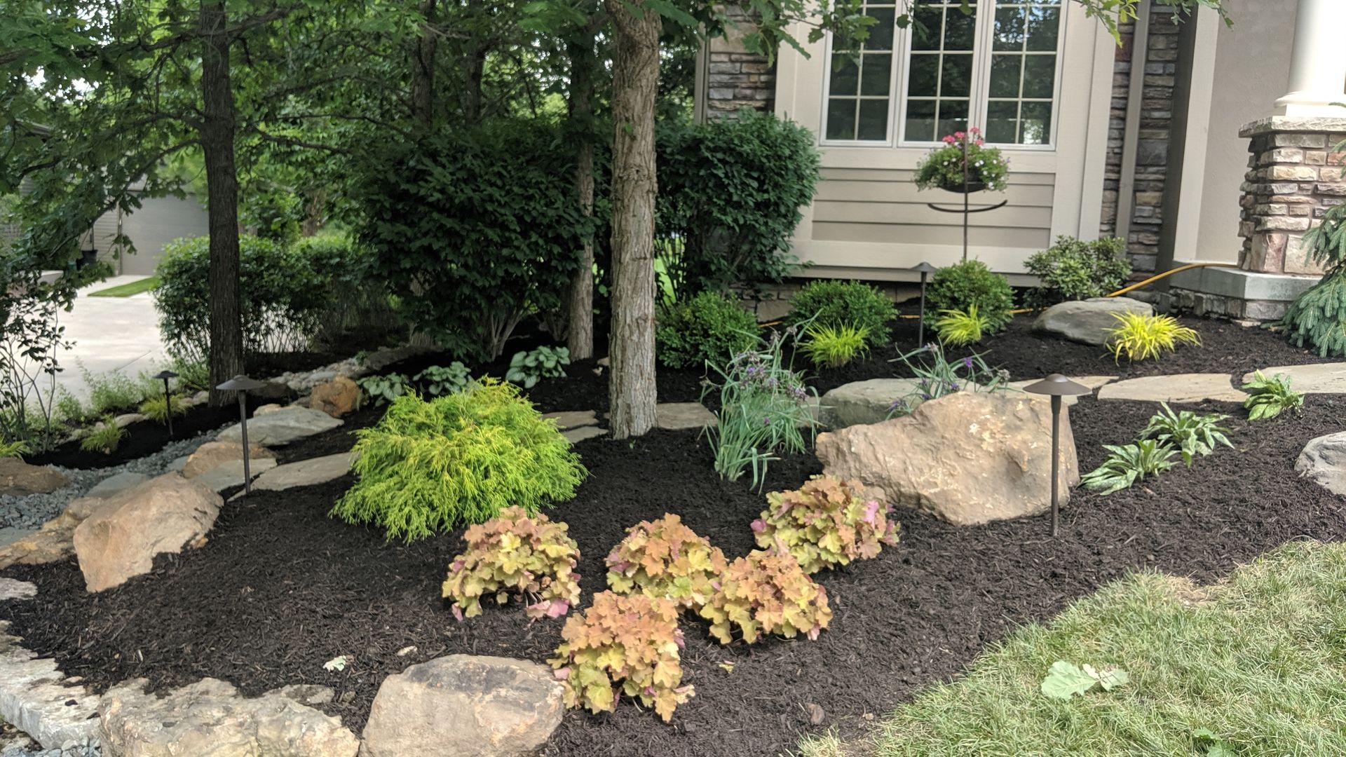 Landscaped front yard with trees, bushes, flowers, rocks, and dark mulch.