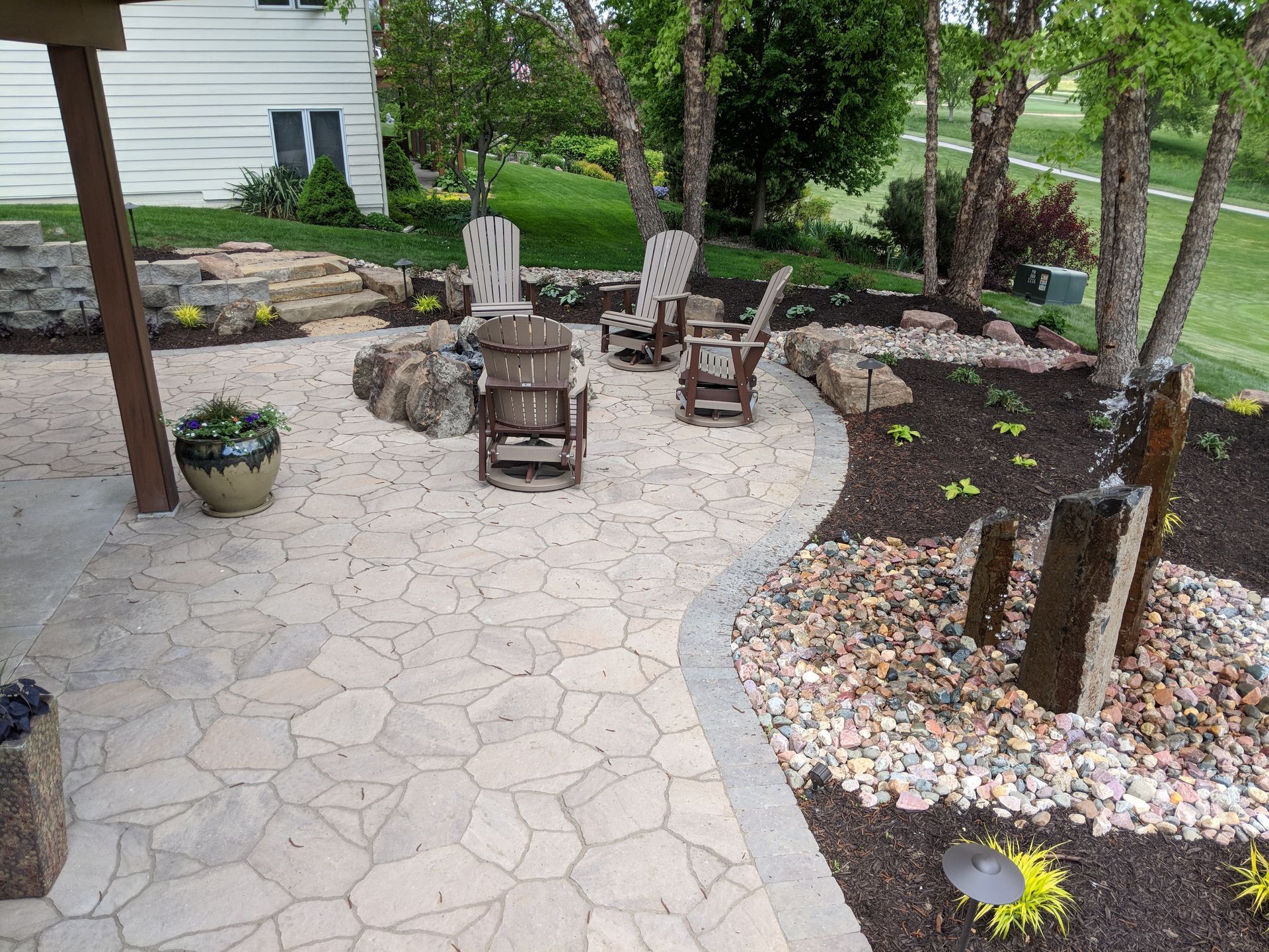 Outdoor patio with stone pavers, seating area, and landscaping with trees and a water feature.