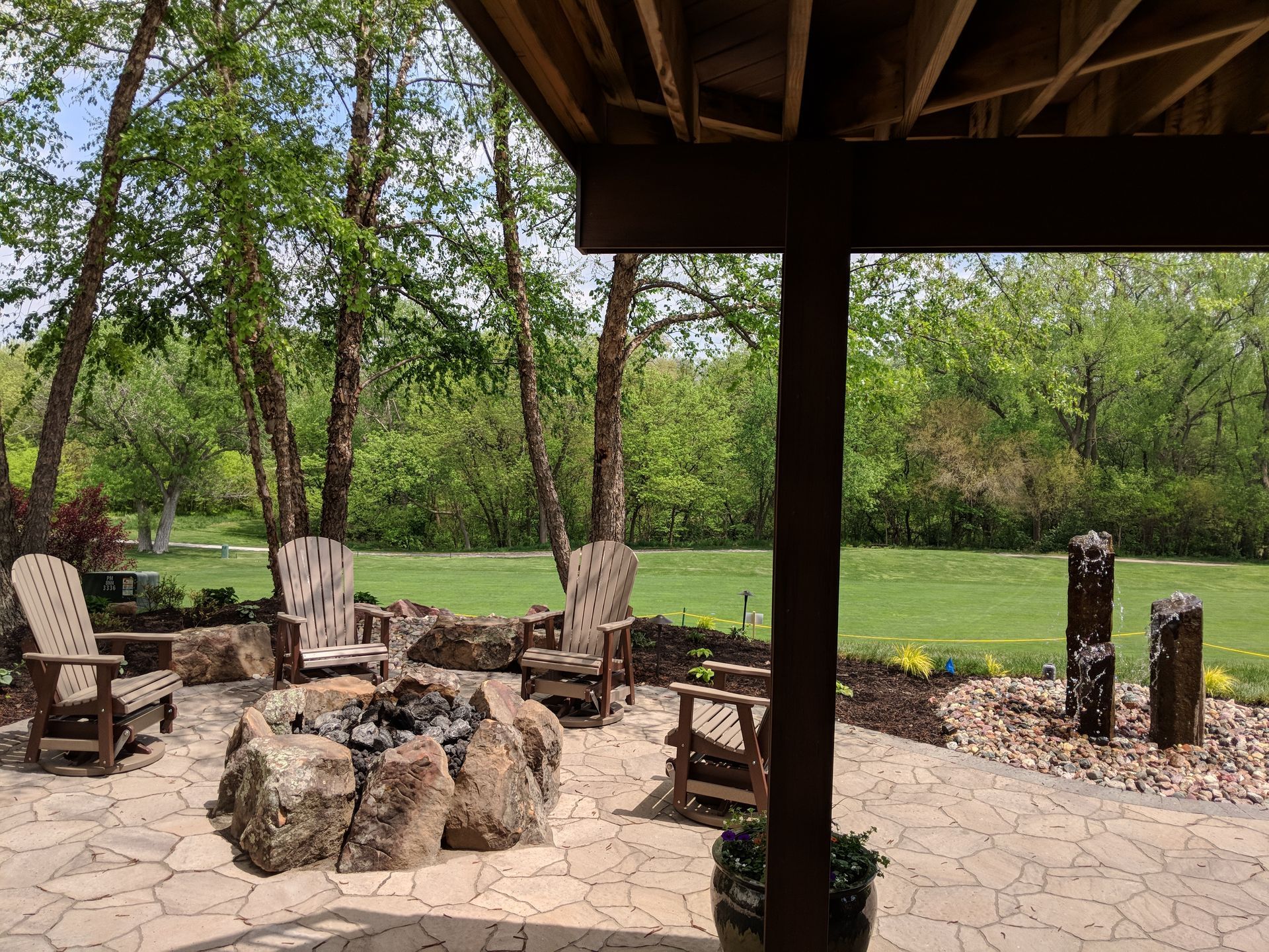 Patio with fire pit, wooden chairs, and fountain under a deck with trees and green grass.