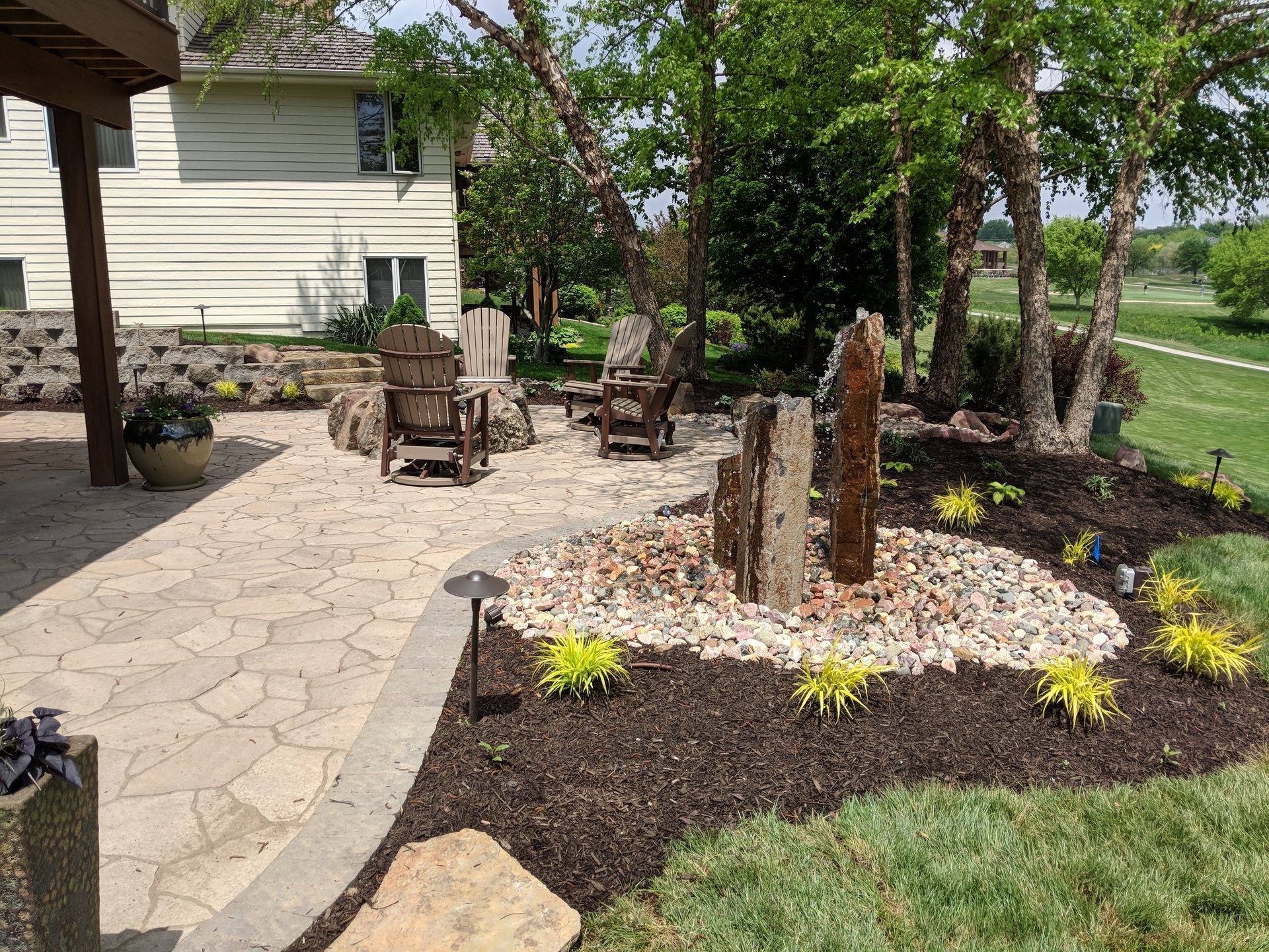 Patio with rock fountain, seating, and landscaping next to a house.