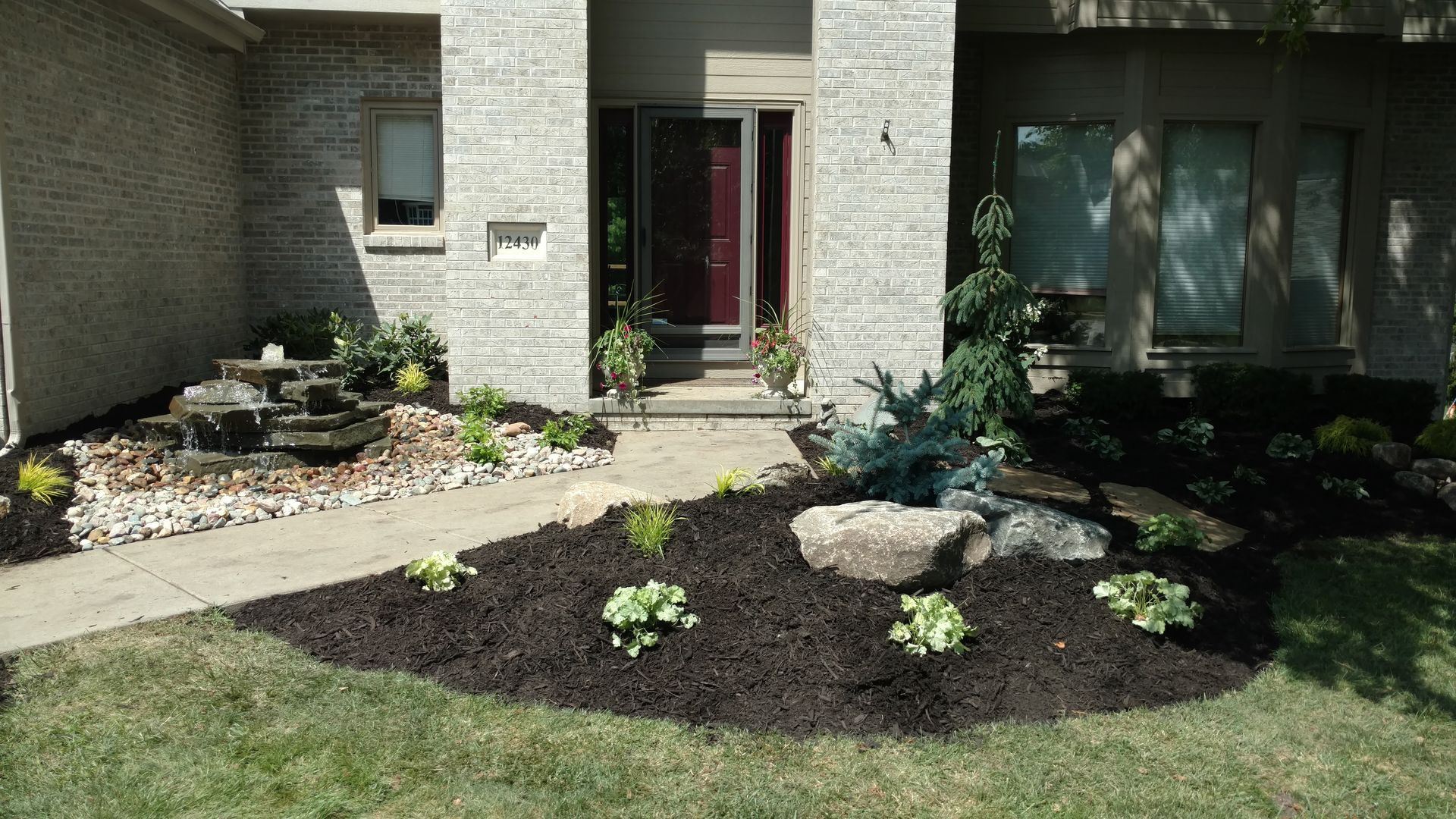 Landscaped front yard with dark mulch, various plants, a water feature, and a stone facade.