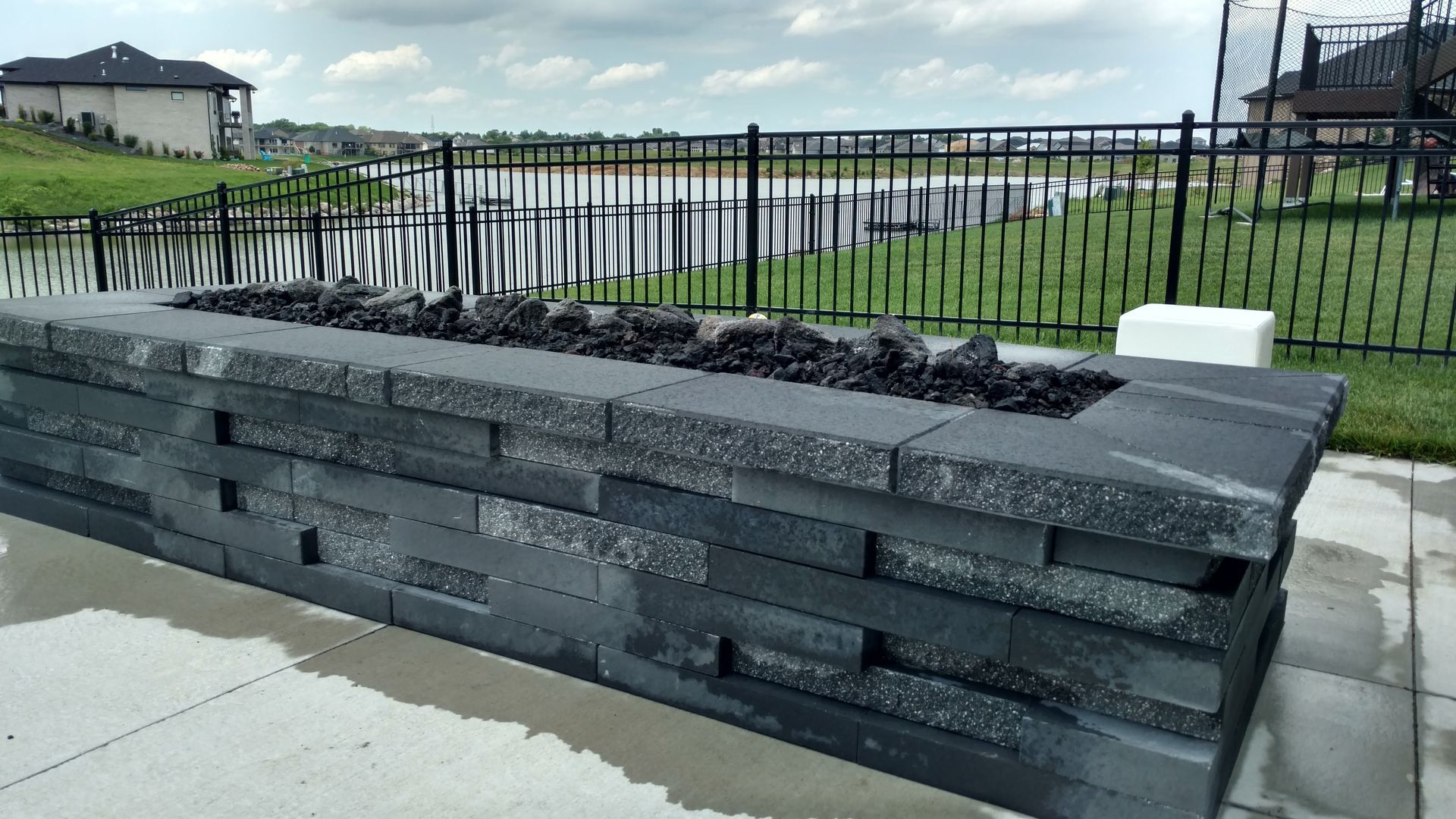 Gray brick fire pit with black glass, outdoor setting near lake and fence.