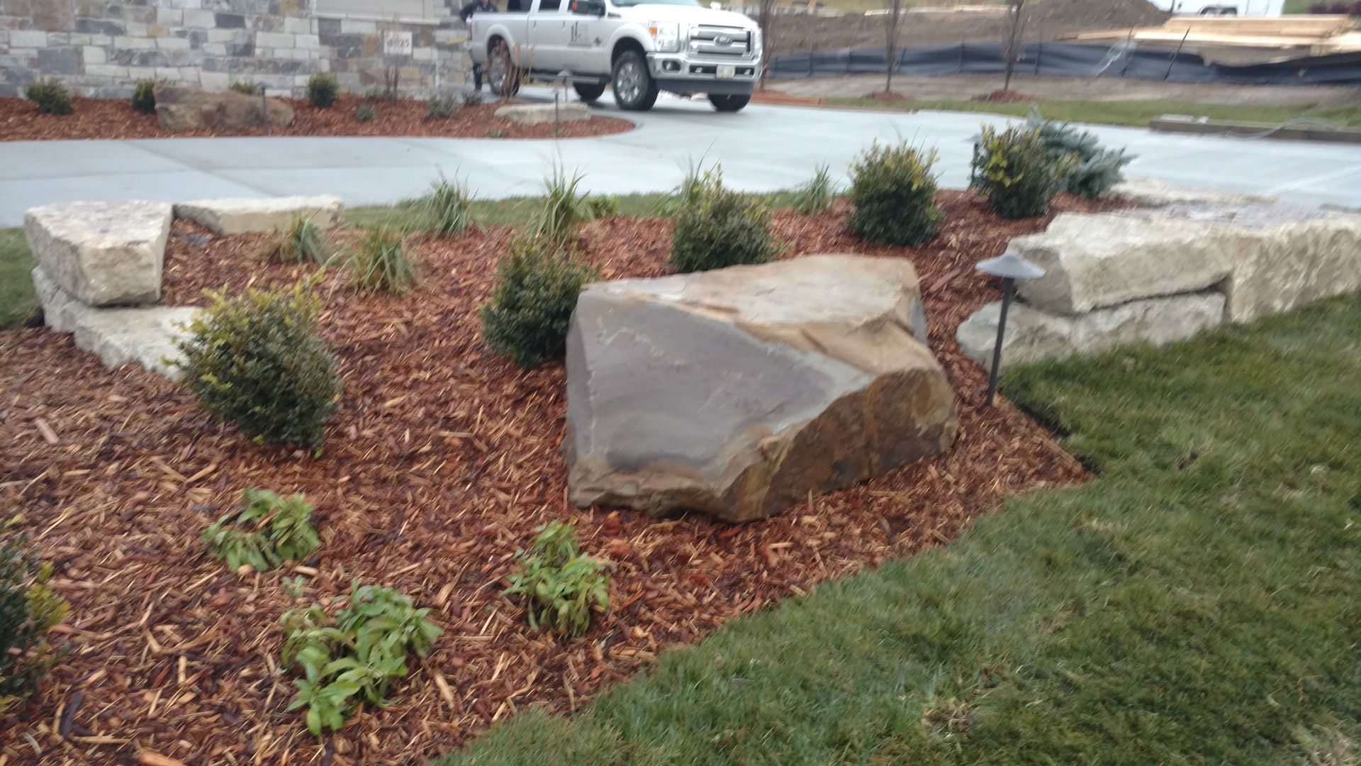 Landscaped garden bed with large rock, mulch, and greenery; truck in background.