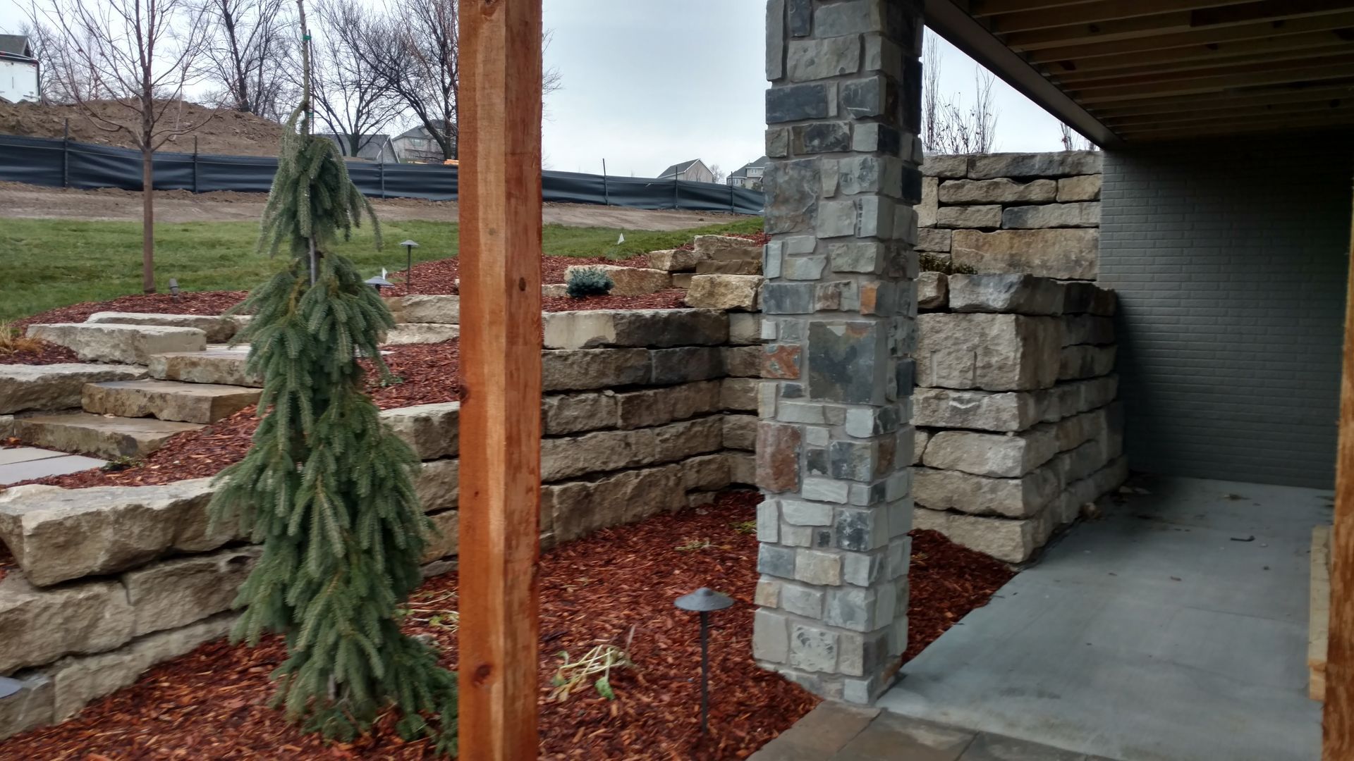 Stone retaining walls and steps with a small evergreen tree. Red mulch. Covered area with stone column.
