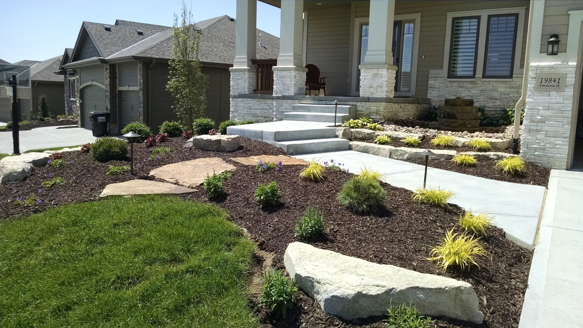 Landscaped front yard with layered stone, mulch, shrubs, and a concrete walkway leading to a house.