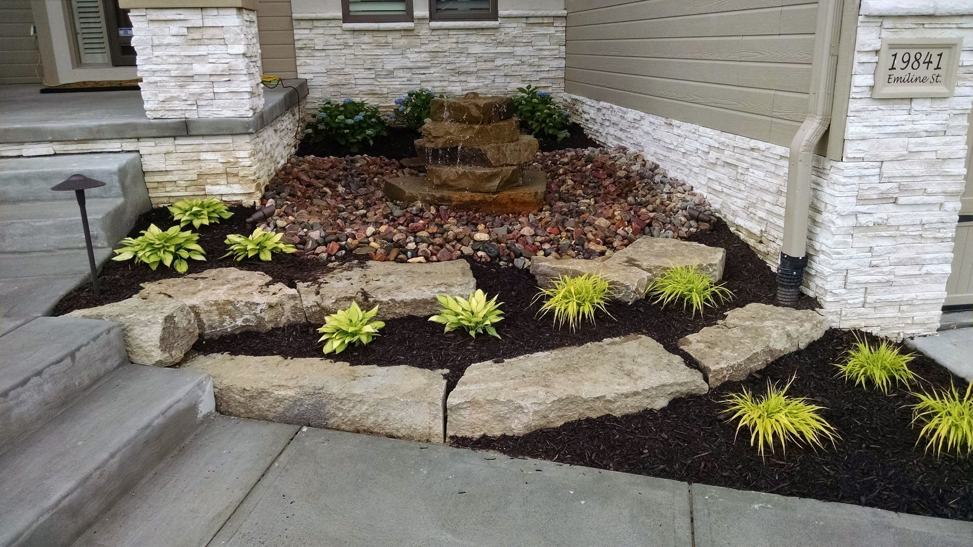 Landscaped yard with stone retaining walls, small fountain, mulch, and green plants.