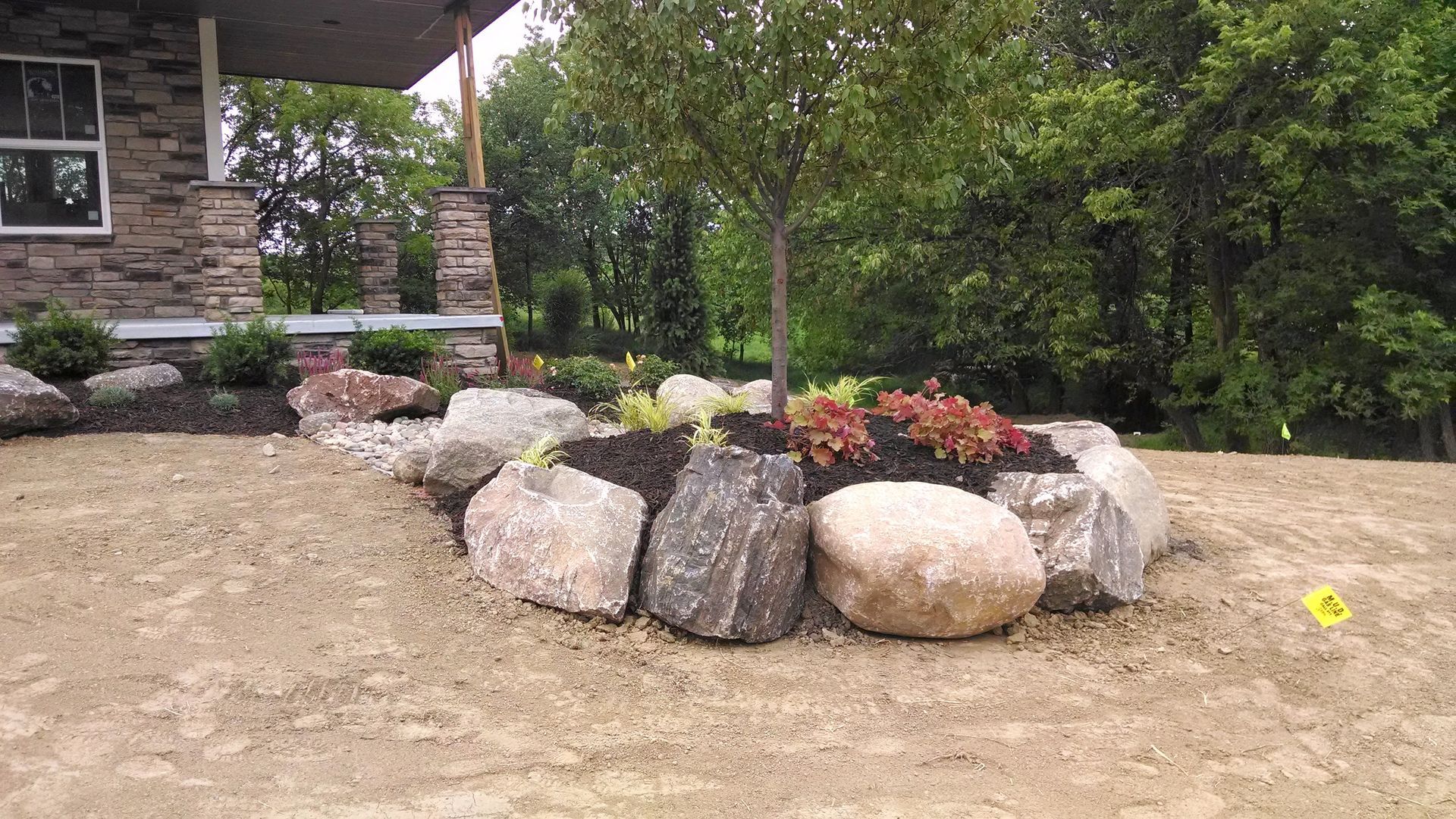 Landscaped front yard with large rocks, a small tree, and colorful plants near a house.