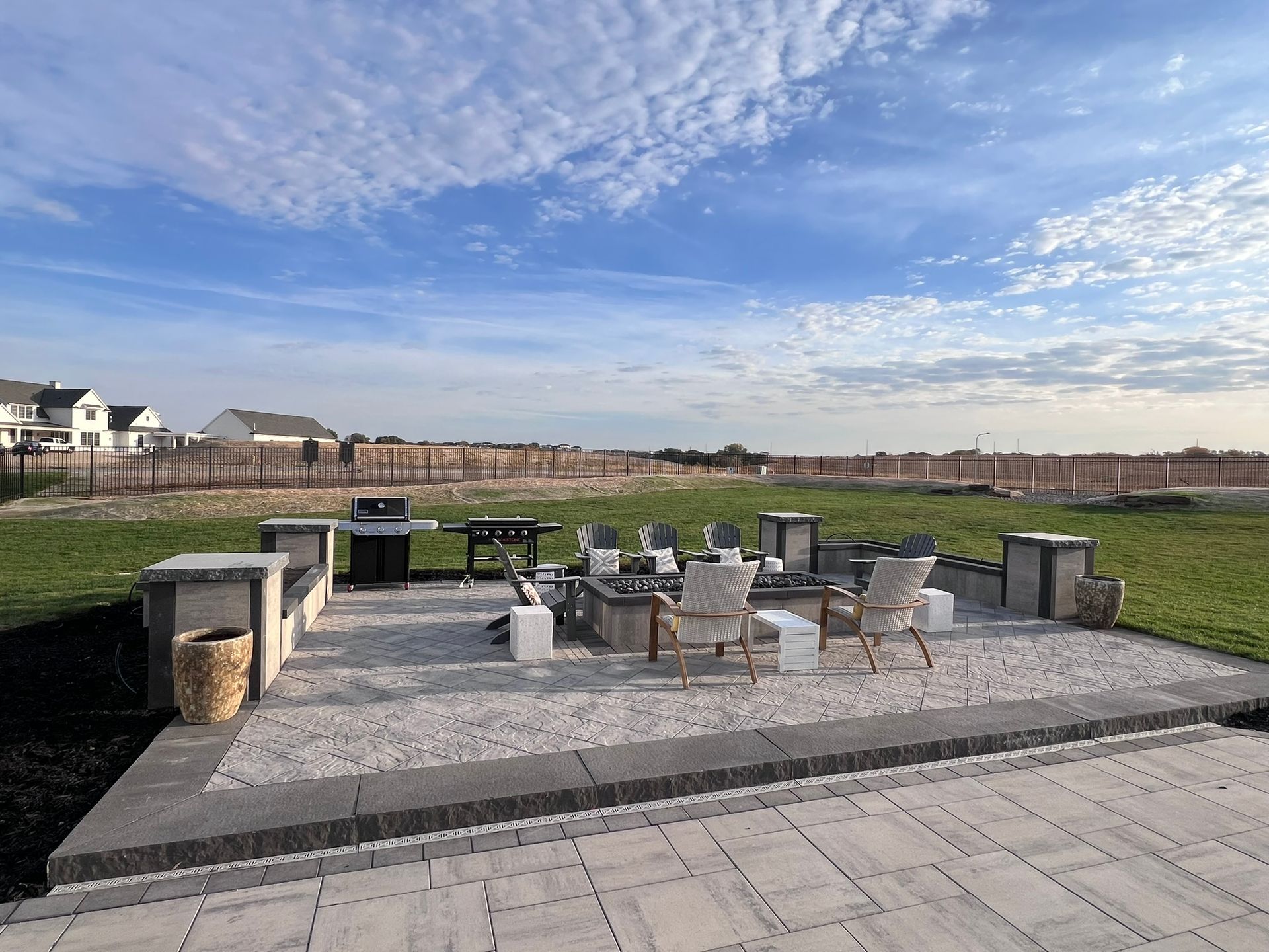 Outdoor patio with seating, grill, and fire pit on a gravel and paved surface under a blue sky.