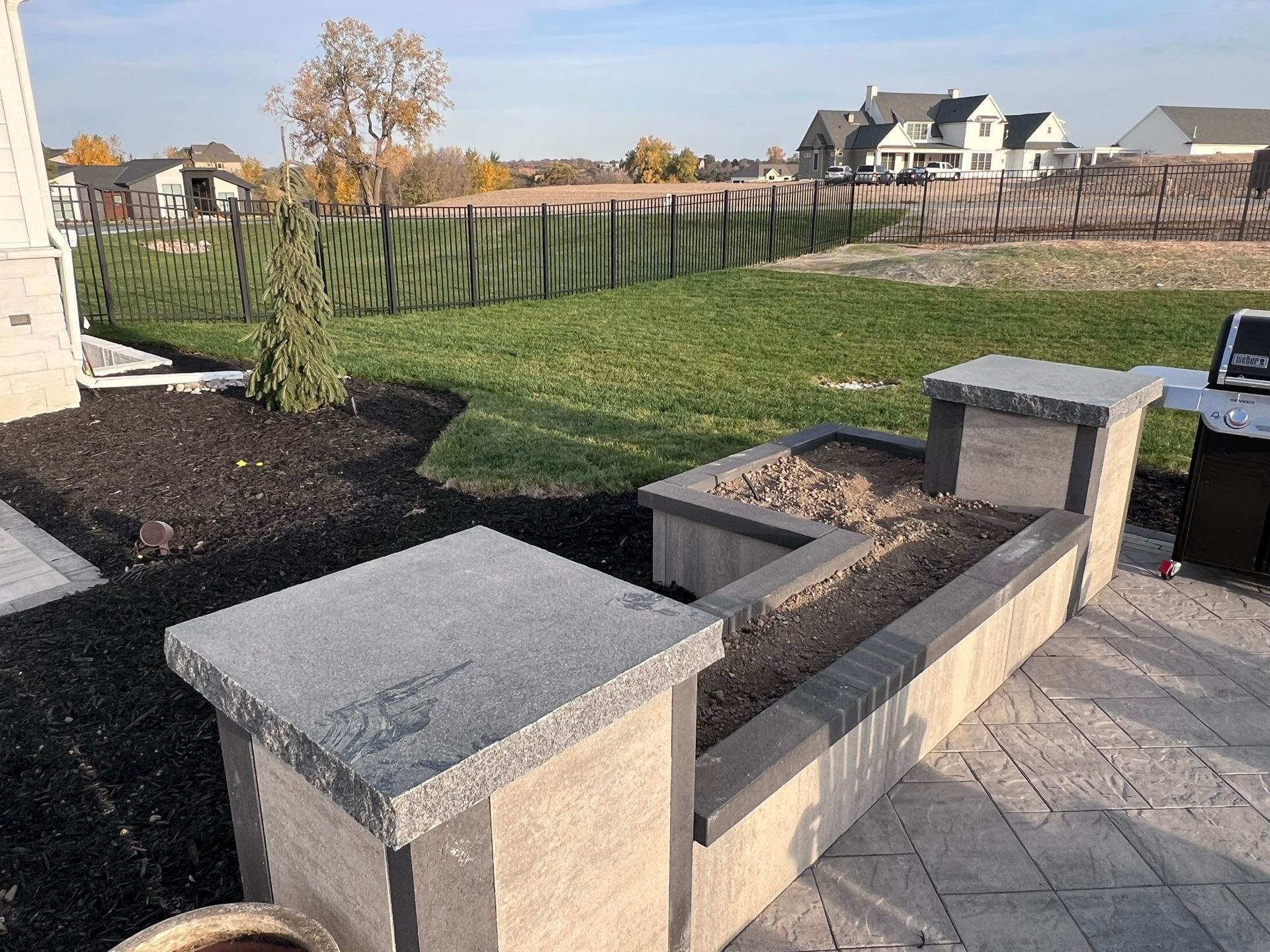 Patio with planter boxes, dark mulch, and a grassy backyard with a fence and houses.
