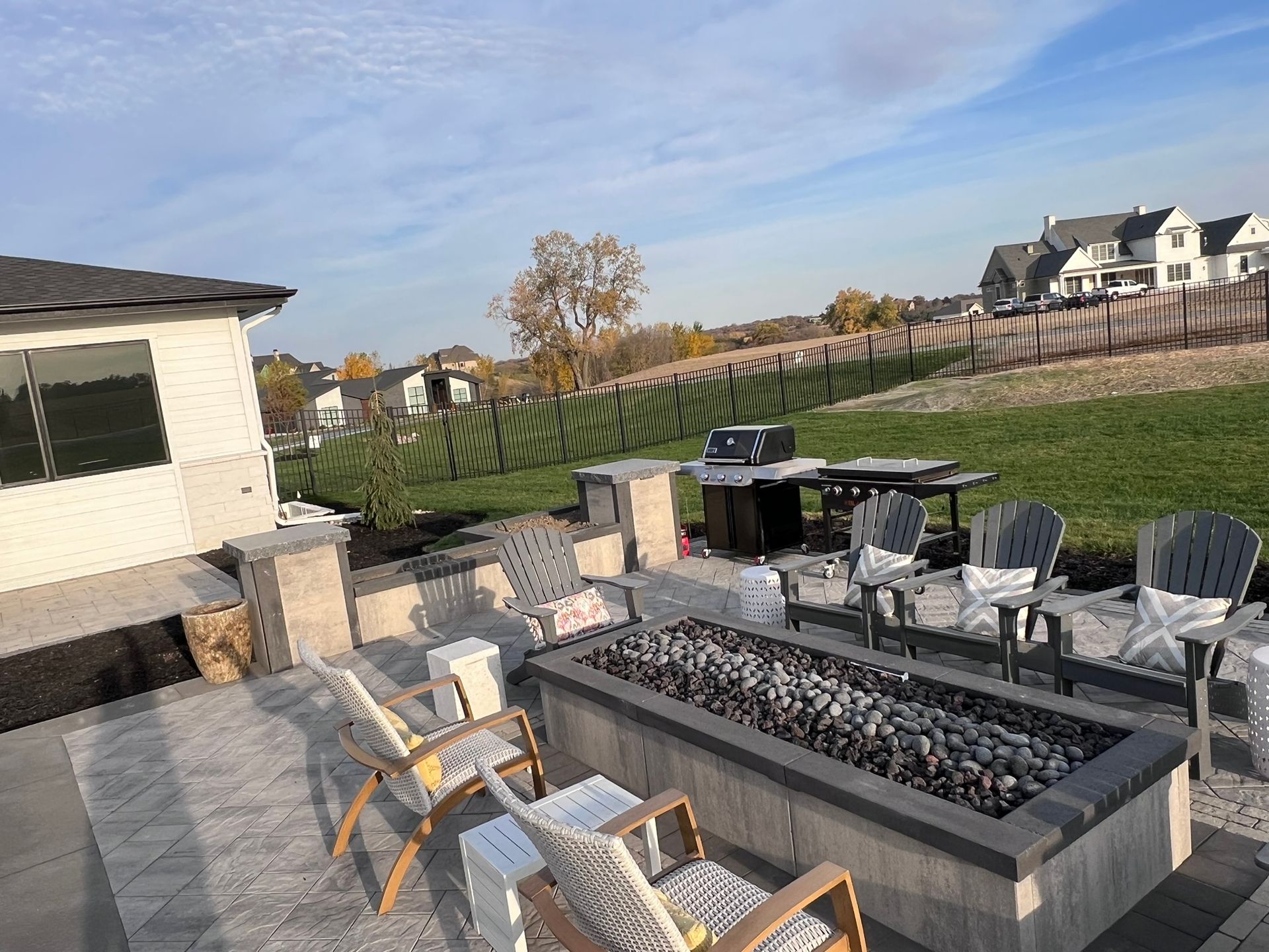 Outdoor patio with fire pit, grill, seating, and a grassy backyard under a blue sky.