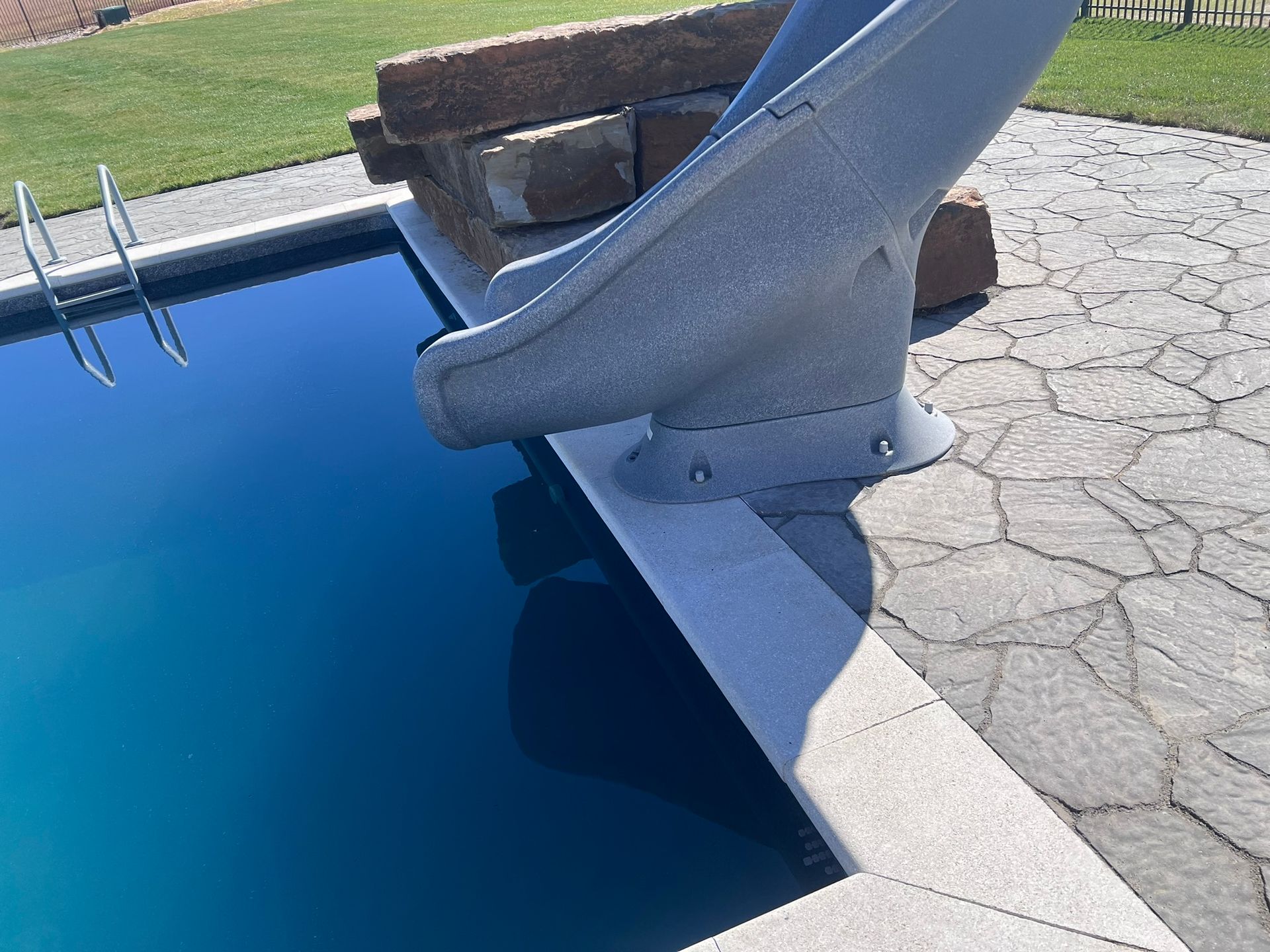 Pool with blue water and a gray slide extending from a stone patio. A ladder is visible.
