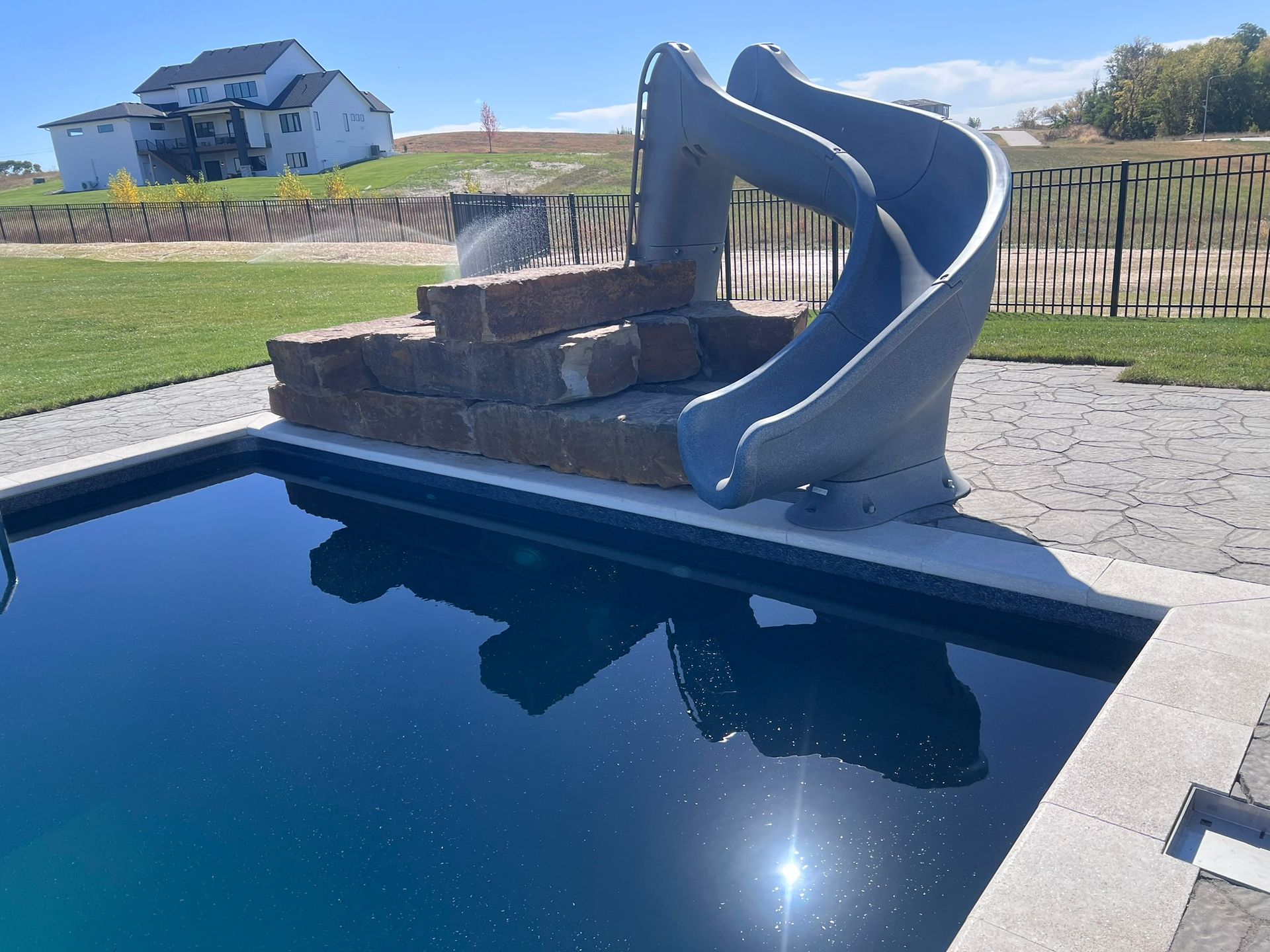Pool with stone steps, a grey slide, and a large house in the background on a sunny day.