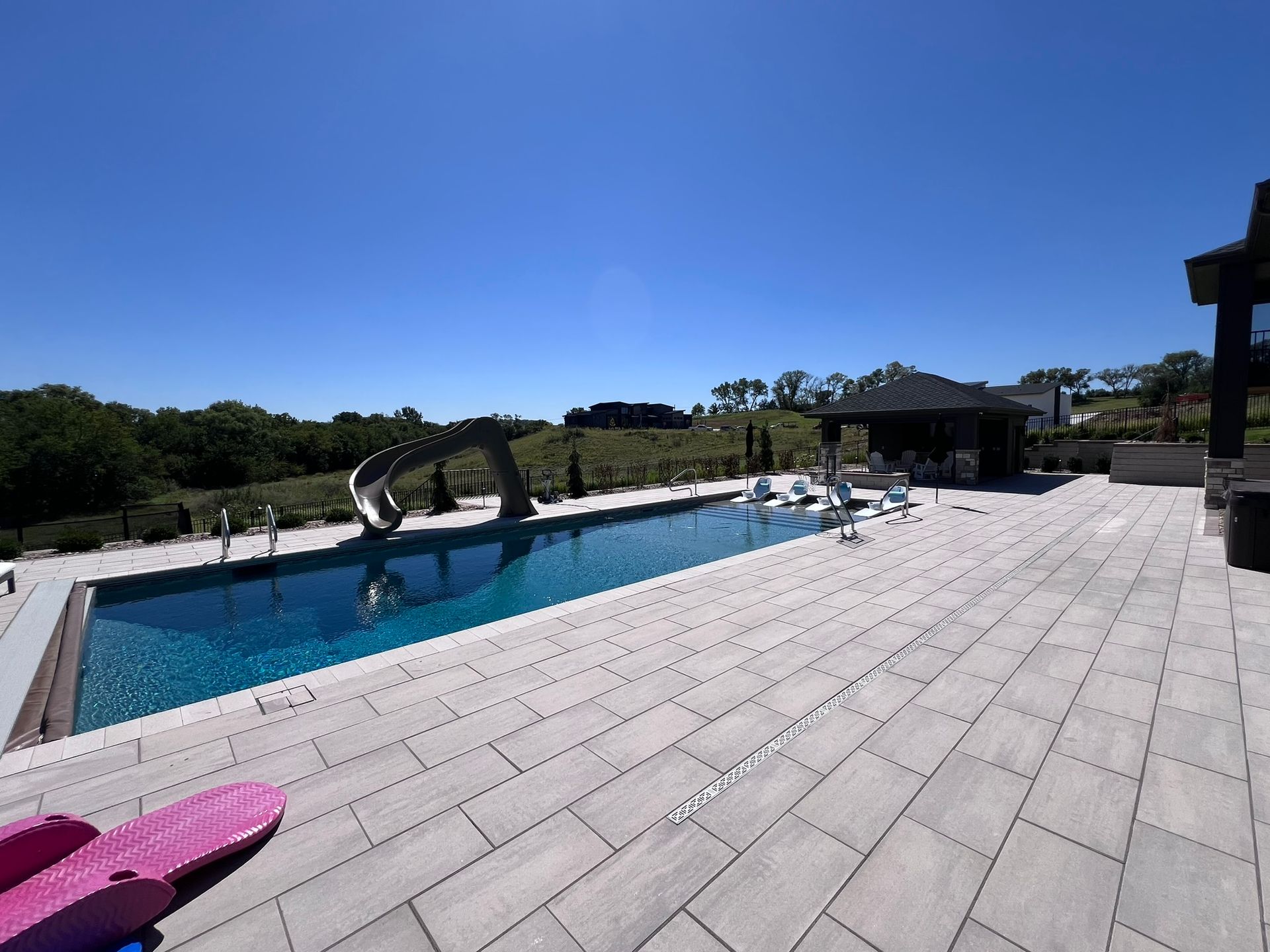 A rectangular pool with a water slide on a paved patio on a sunny day.