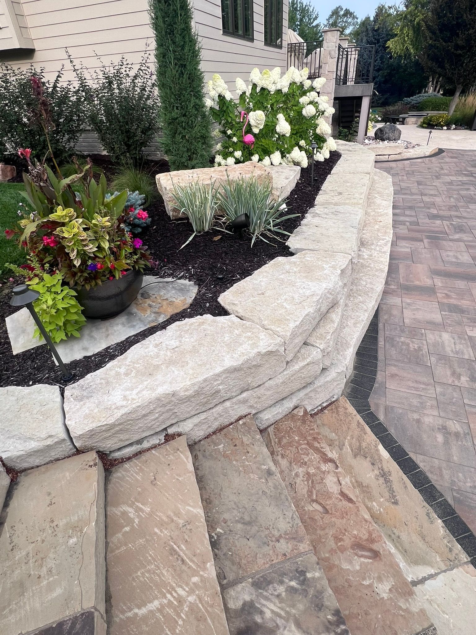 Stone retaining wall with a garden bed and steps, next to a brick building and a paved driveway.