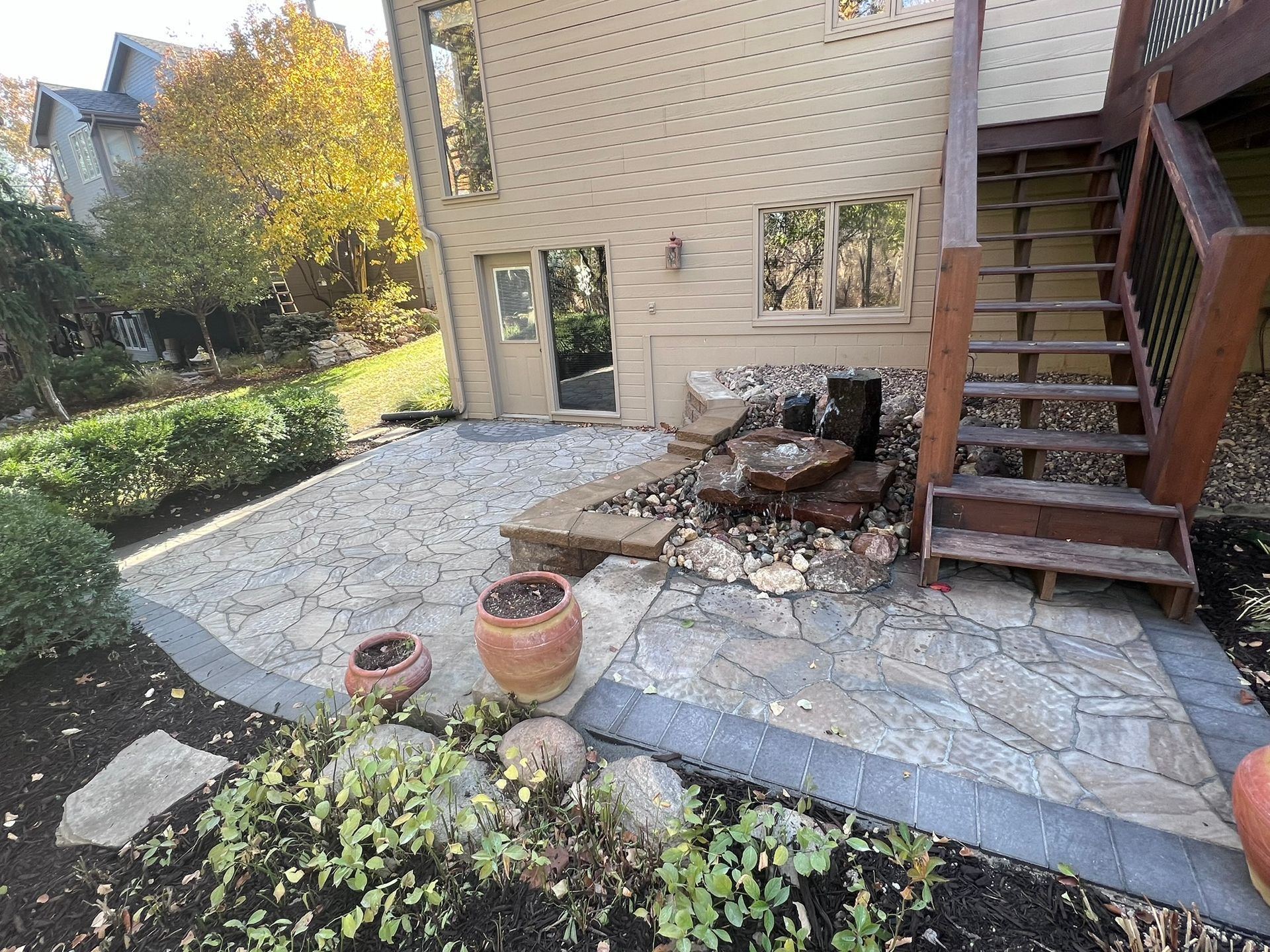 Stone patio with a waterfall feature, next to a house with wooden stairs and landscaping.