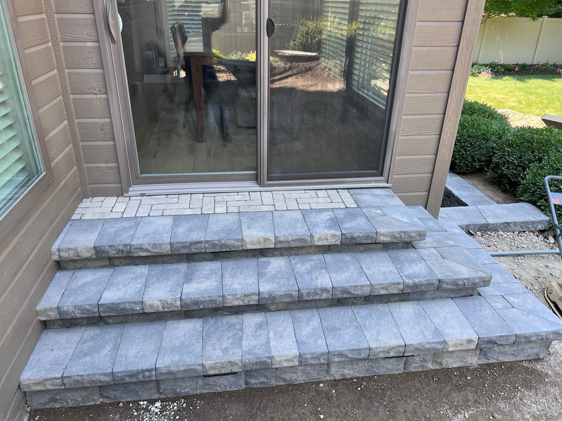 Three-step gray brick staircase leading to sliding glass door. Brick patio with landscaping visible.