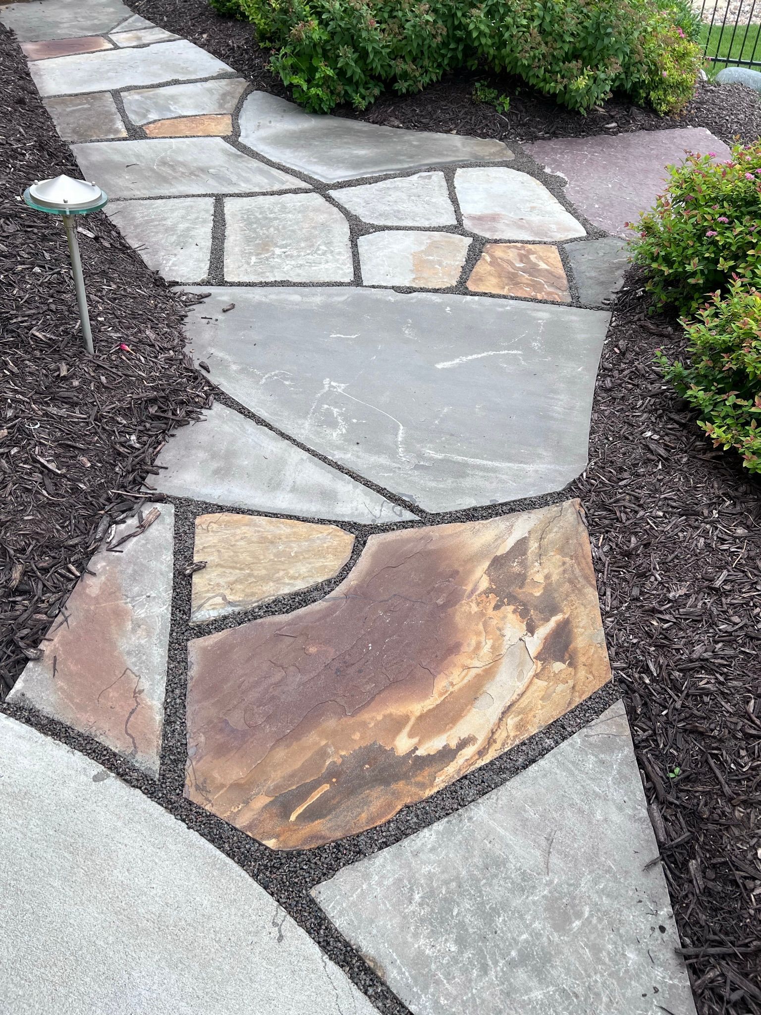Flagstone walkway through a garden, edged with mulch and bushes.