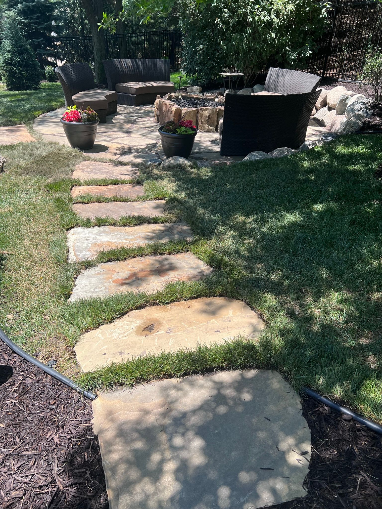 Stone pathway through grass leads to a patio with seating and potted flowers.