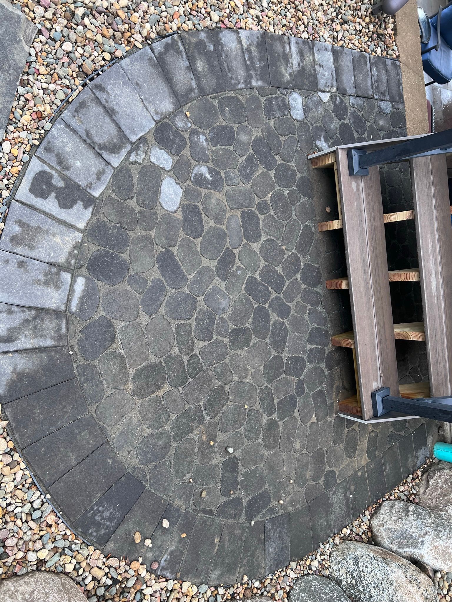 Circular stone patio with dark pavers and edging, next to wooden stairs.
