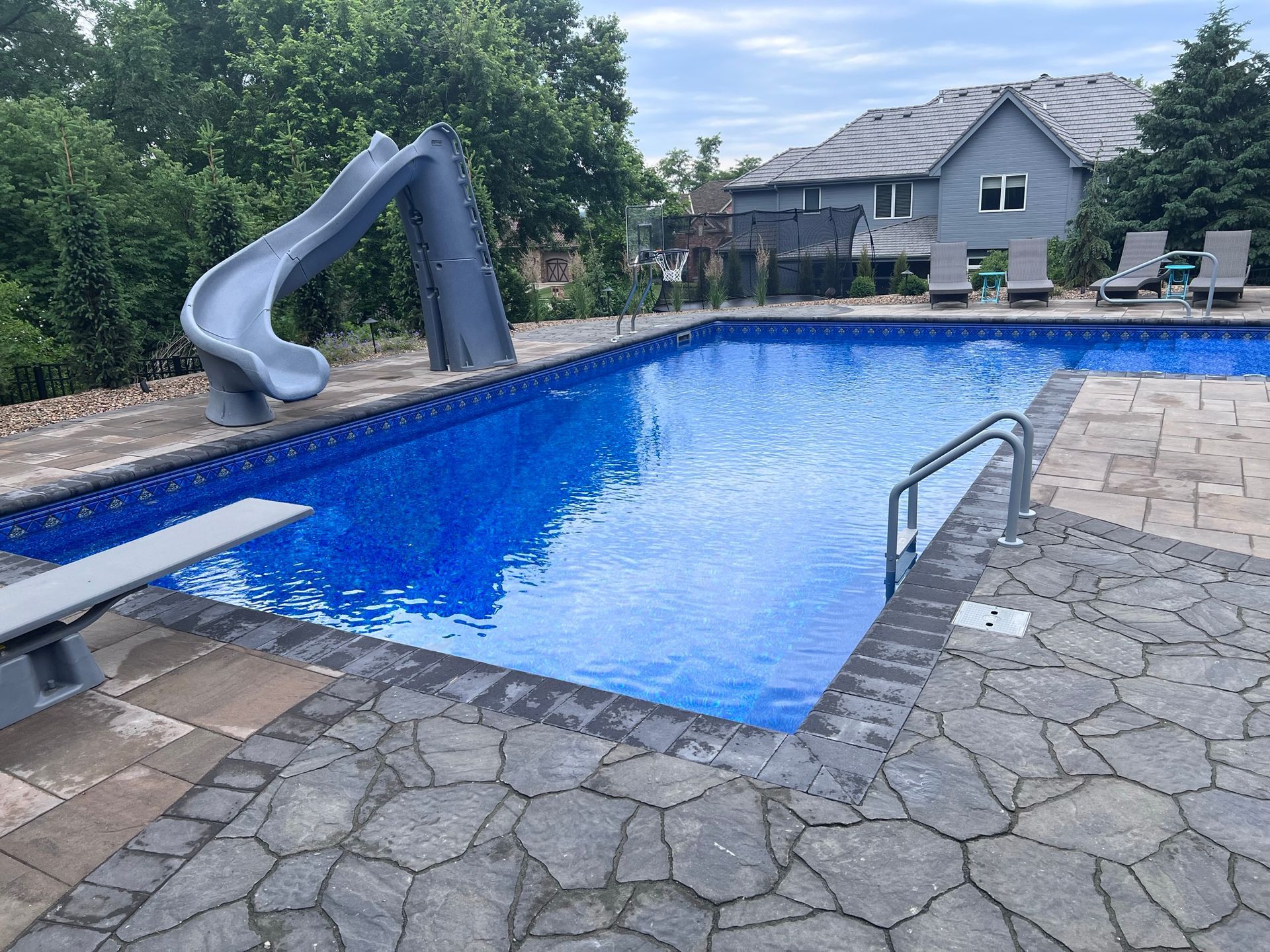 Backyard pool with a slide, diving board, and stone patio. The pool has blue water.