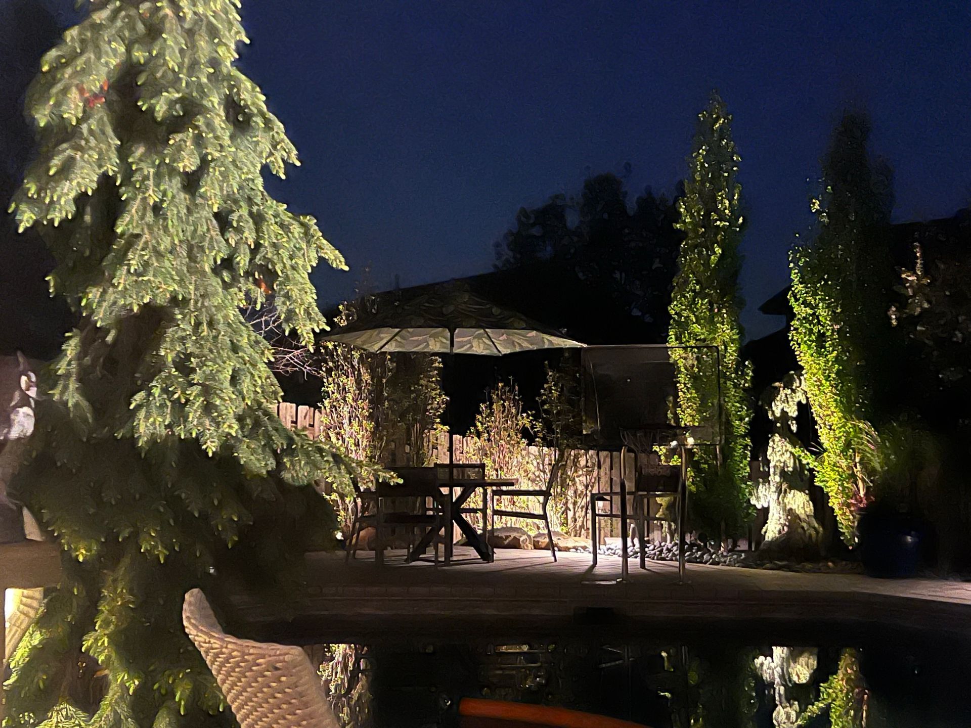 Backyard scene at night with pool, patio table, illuminated trees and gazebo.