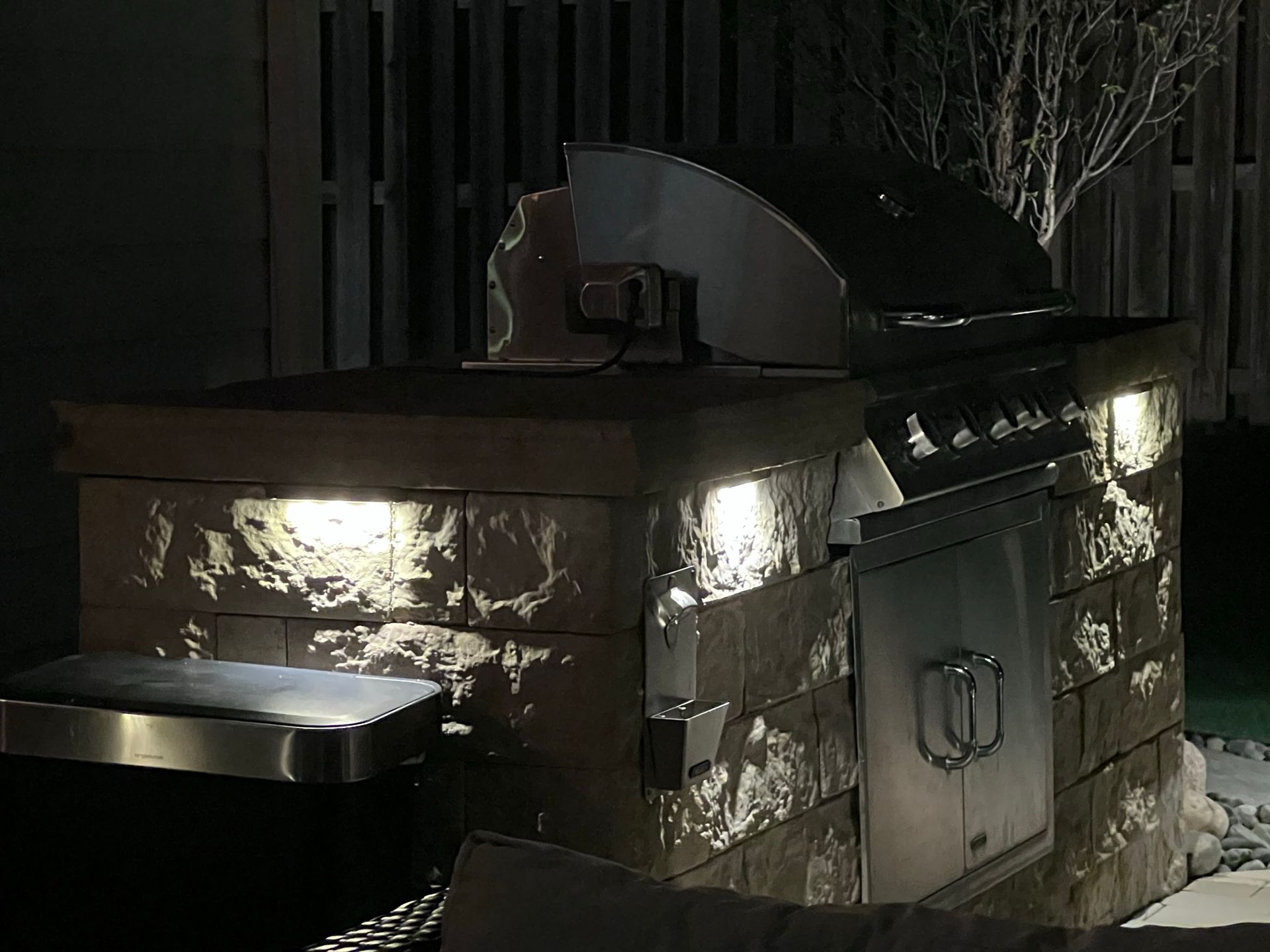 Outdoor grill built into stone structure, illuminated by overhead lights, at night.