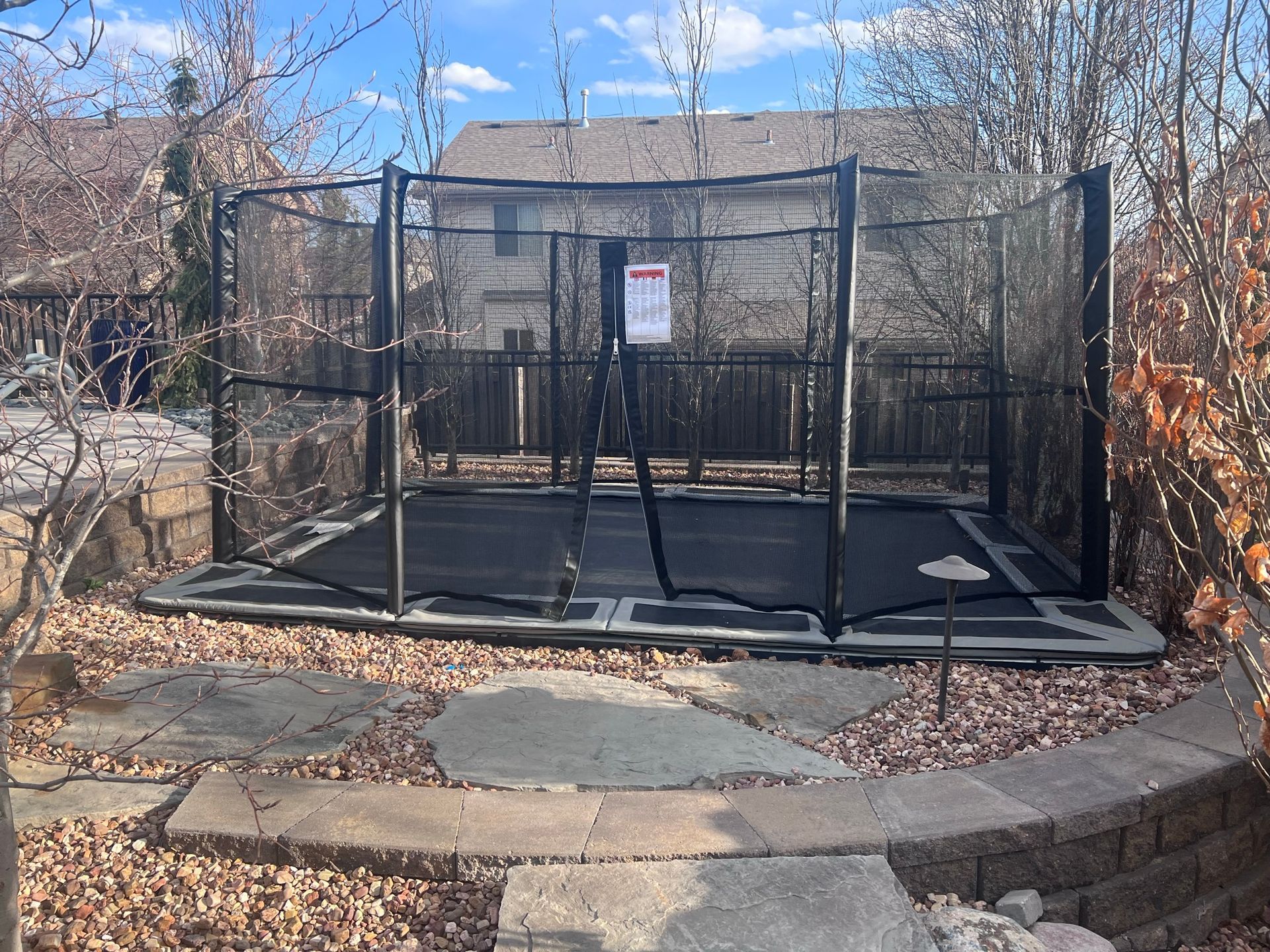 Trampoline with safety net in backyard, surrounded by landscaping and fence.