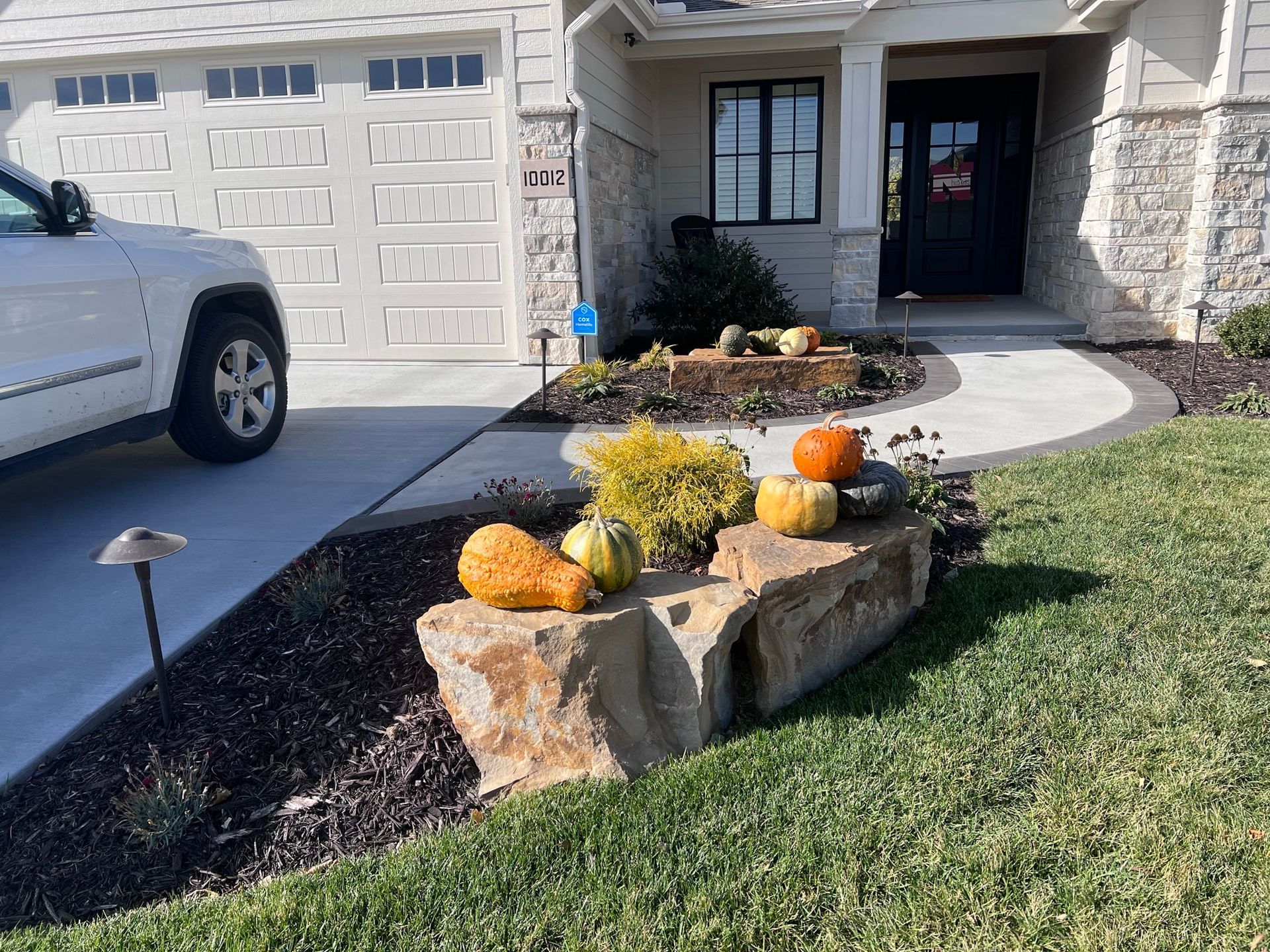 A stone planter with pumpkins and gourds near a house's entryway and driveway.