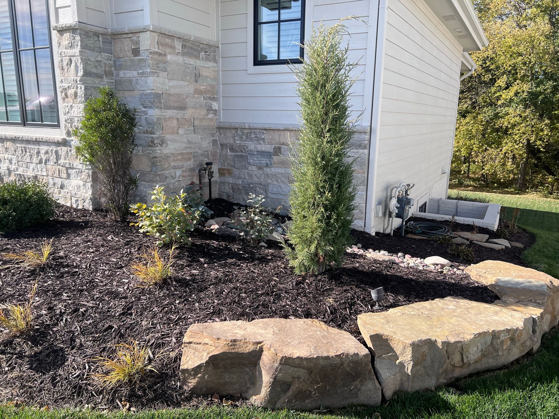 Exterior of a house with stone and white siding, landscaped with mulch, plants, and large rock border.