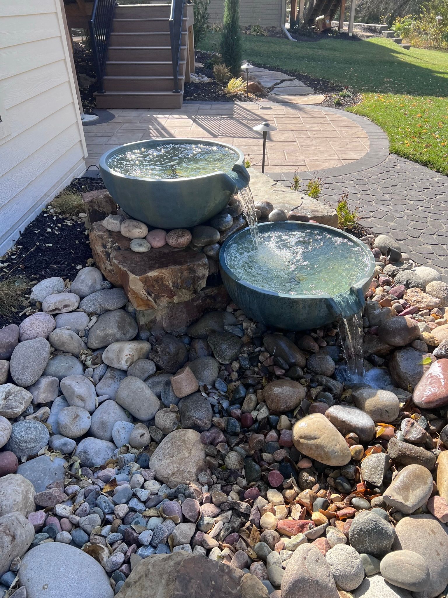 Water fountain with two turquoise bowls, rocks, and a brick pathway in a garden setting.