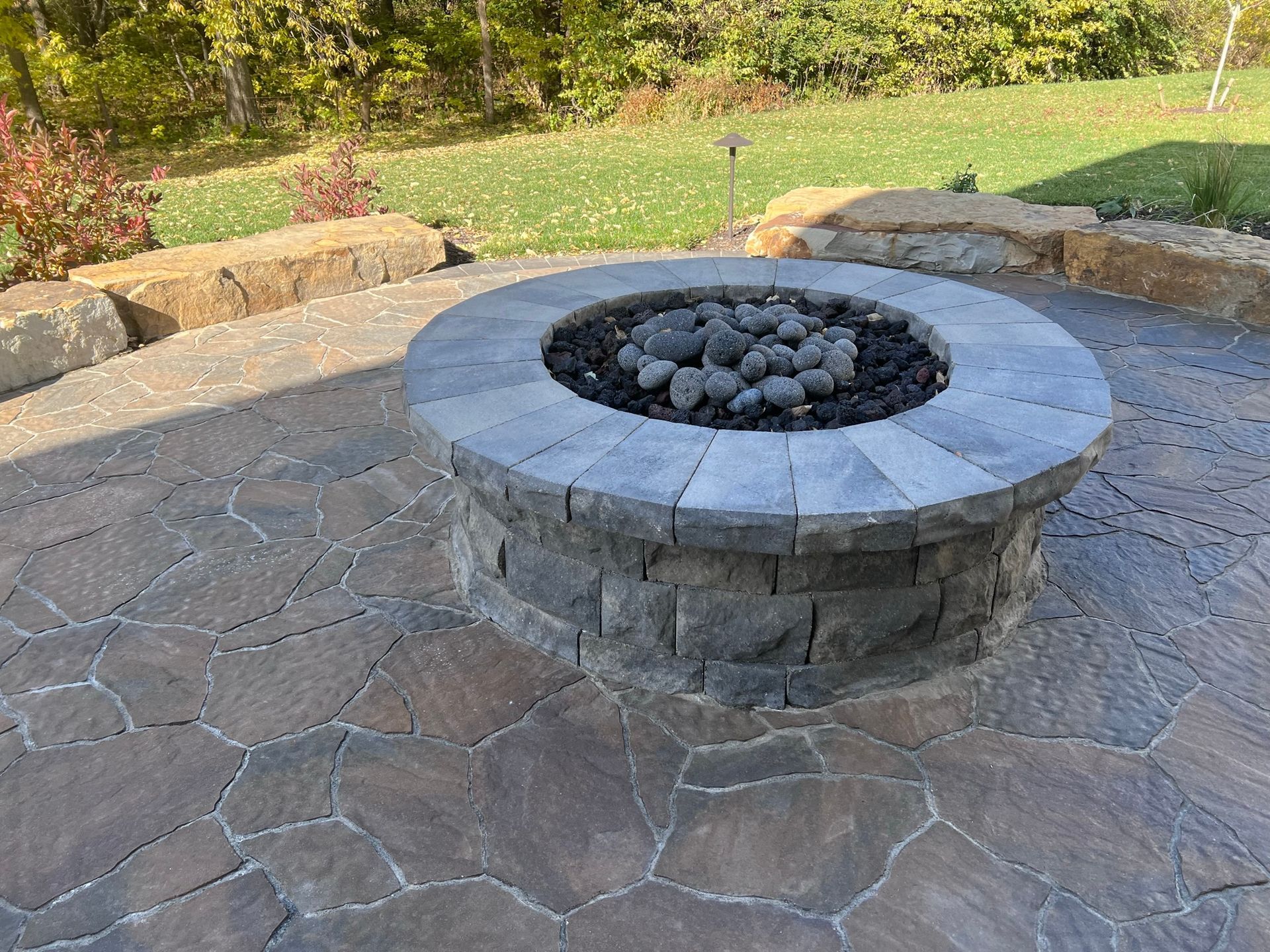 Circular stone fire pit on a patterned stone patio, surrounded by a grassy lawn and some landscaping.