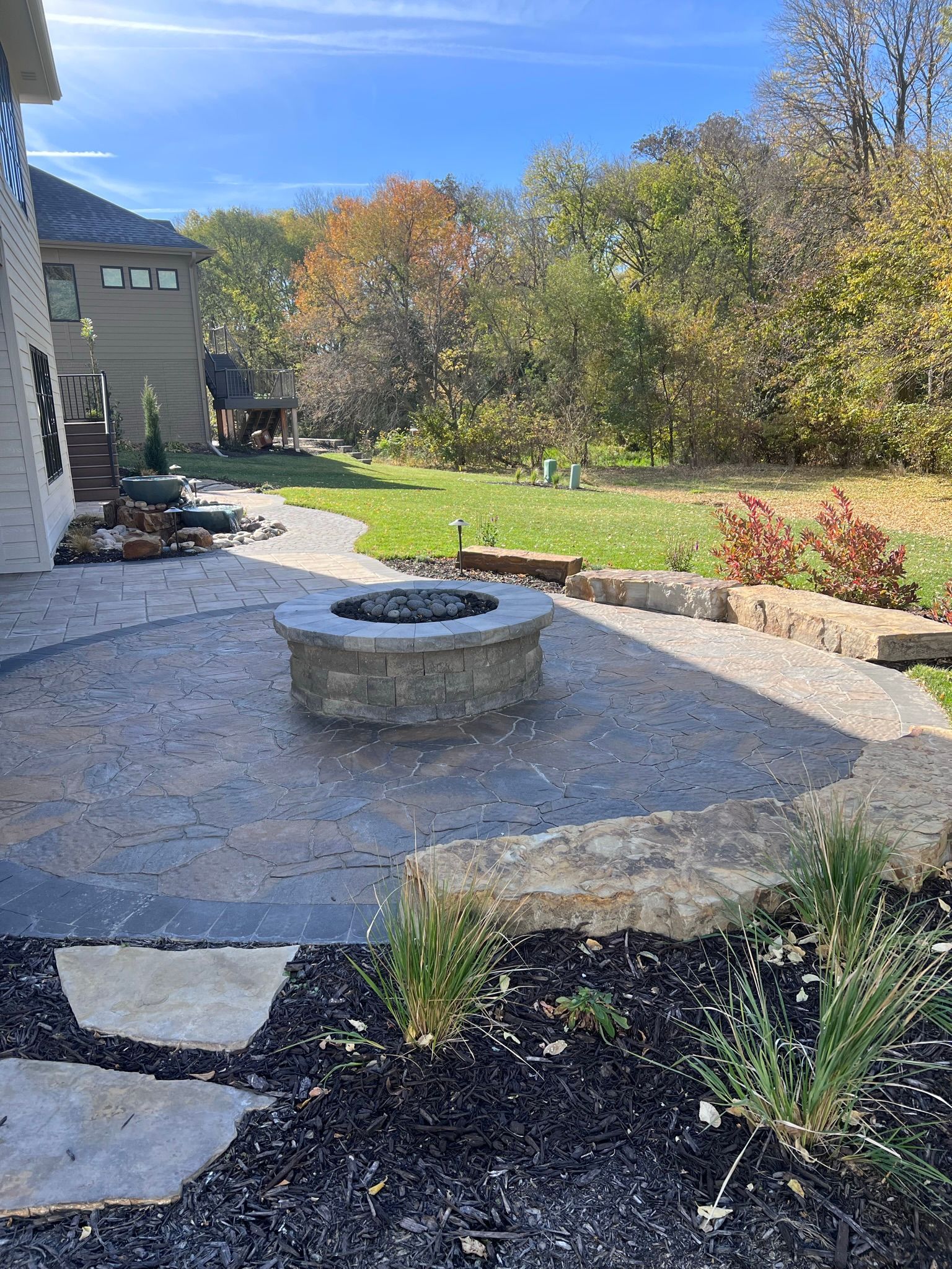 Backyard patio with fire pit, bordering lawn and wooded area under a blue sky.