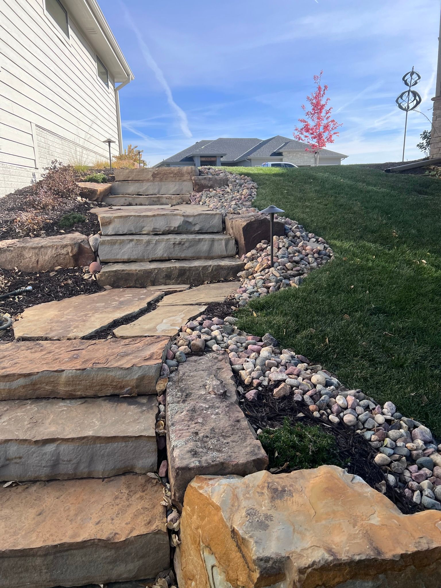 Stone steps ascend a sloped, landscaped yard. A building is to the left, and a grassy hill is to the right.