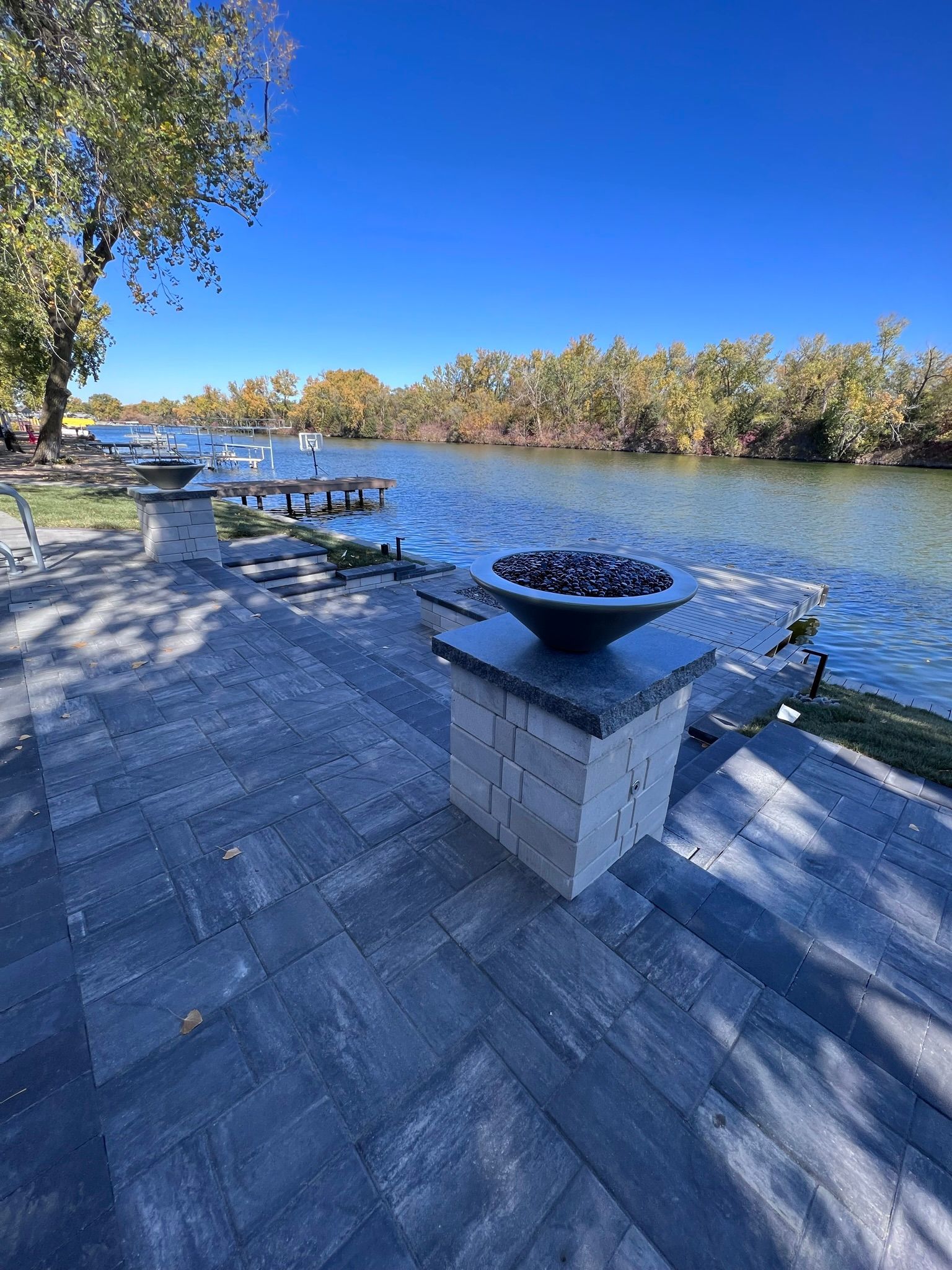 Patio overlooking a river with a fire pit, boats, and autumn trees under a blue sky.