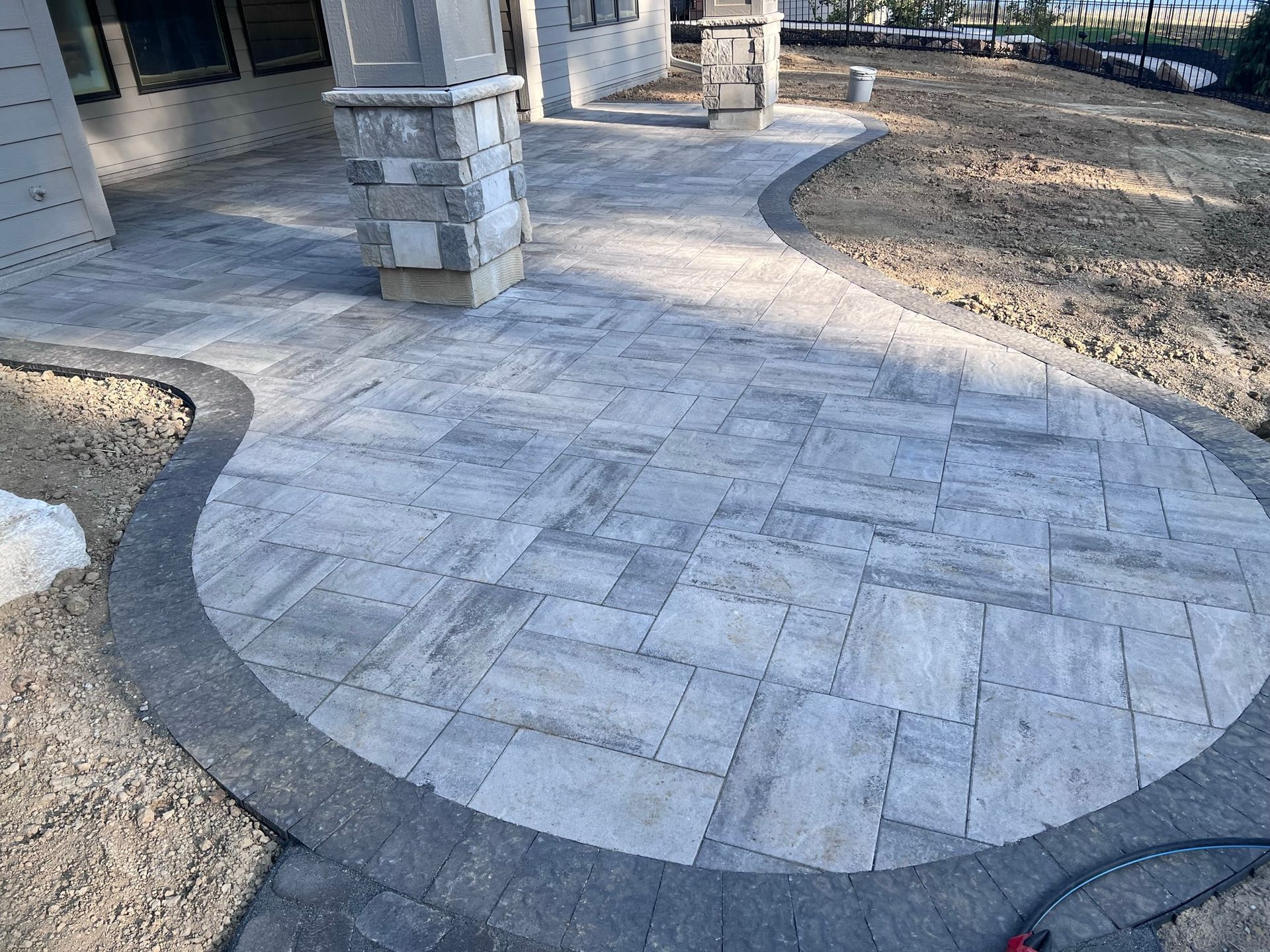 Stone patio with gray pavers and a dark border, near a house with columns and dirt.