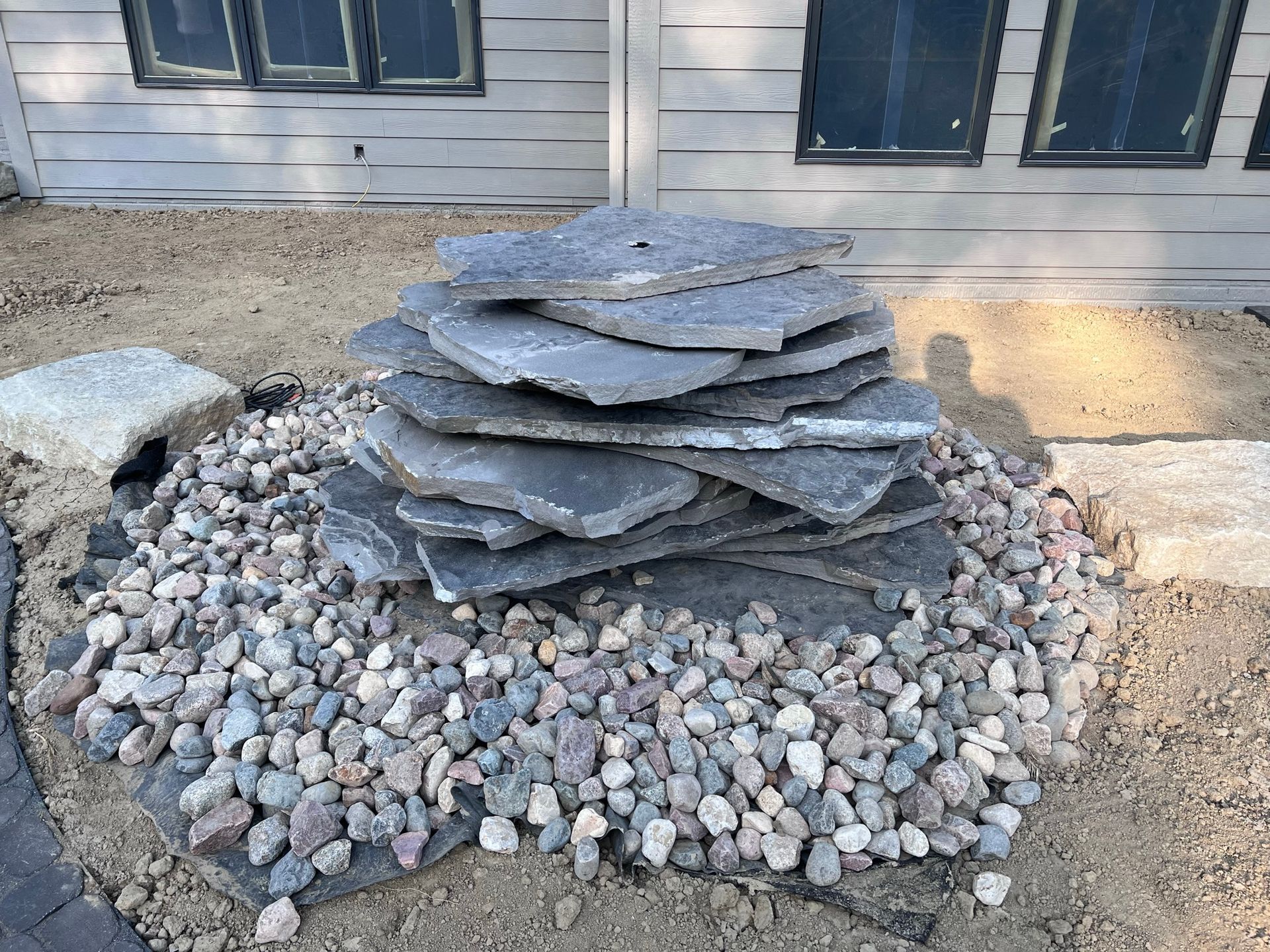 A stacked stone water feature with gravel base, set outdoors in front of a house.