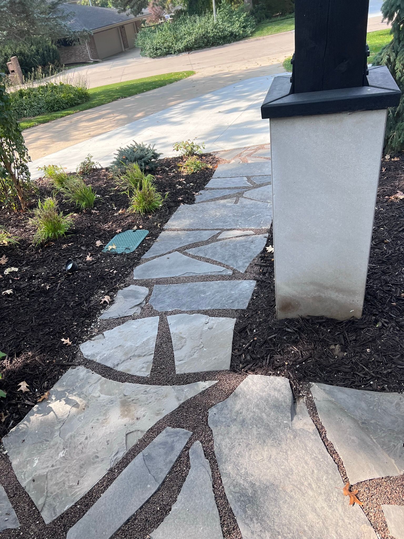 Flagstone pathway next to a white pillar, surrounded by dark mulch and plants; a driveway is in the background.