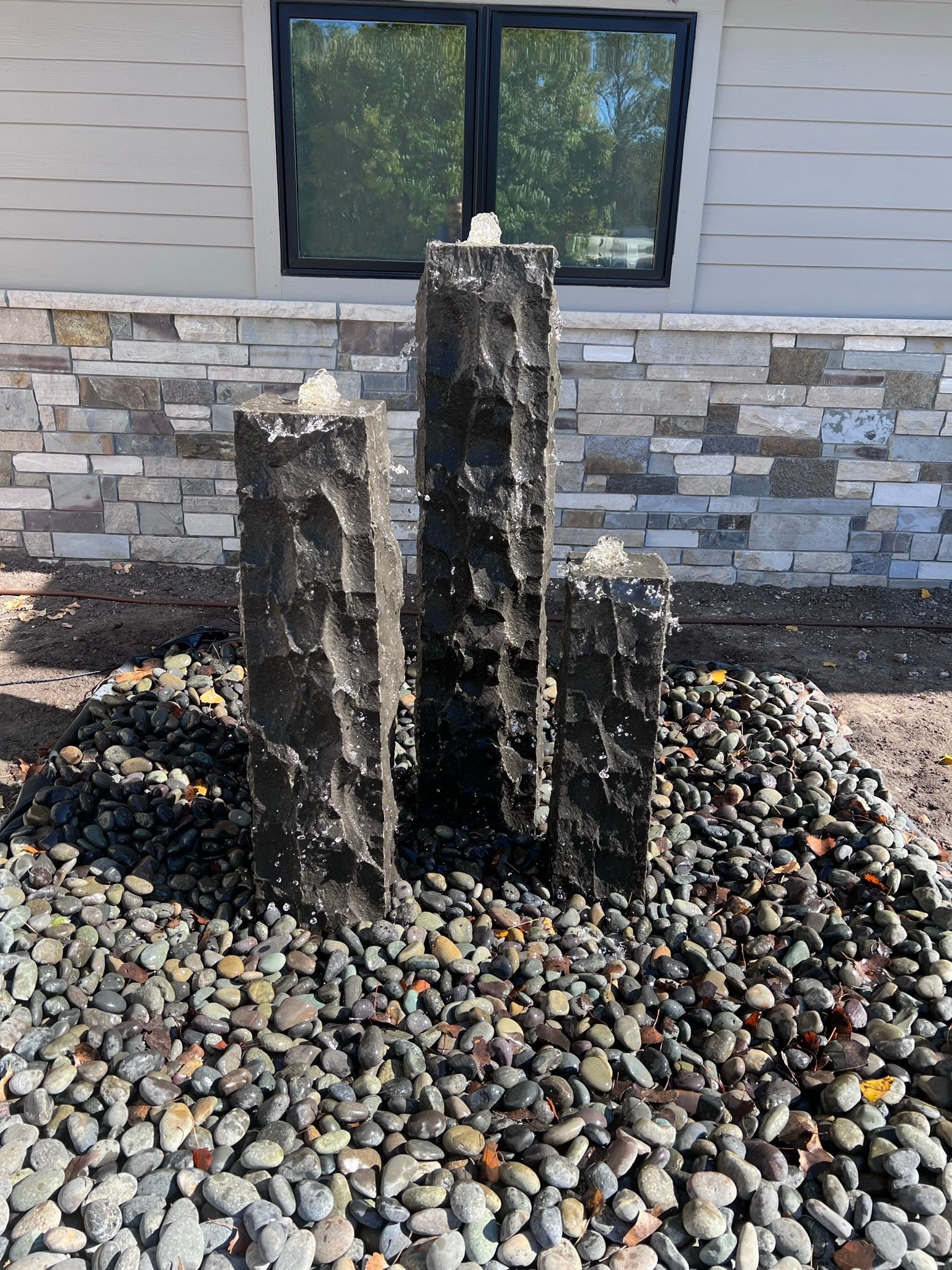 Three black stone water fountains in a bed of pebbles, in front of a house with stone siding.