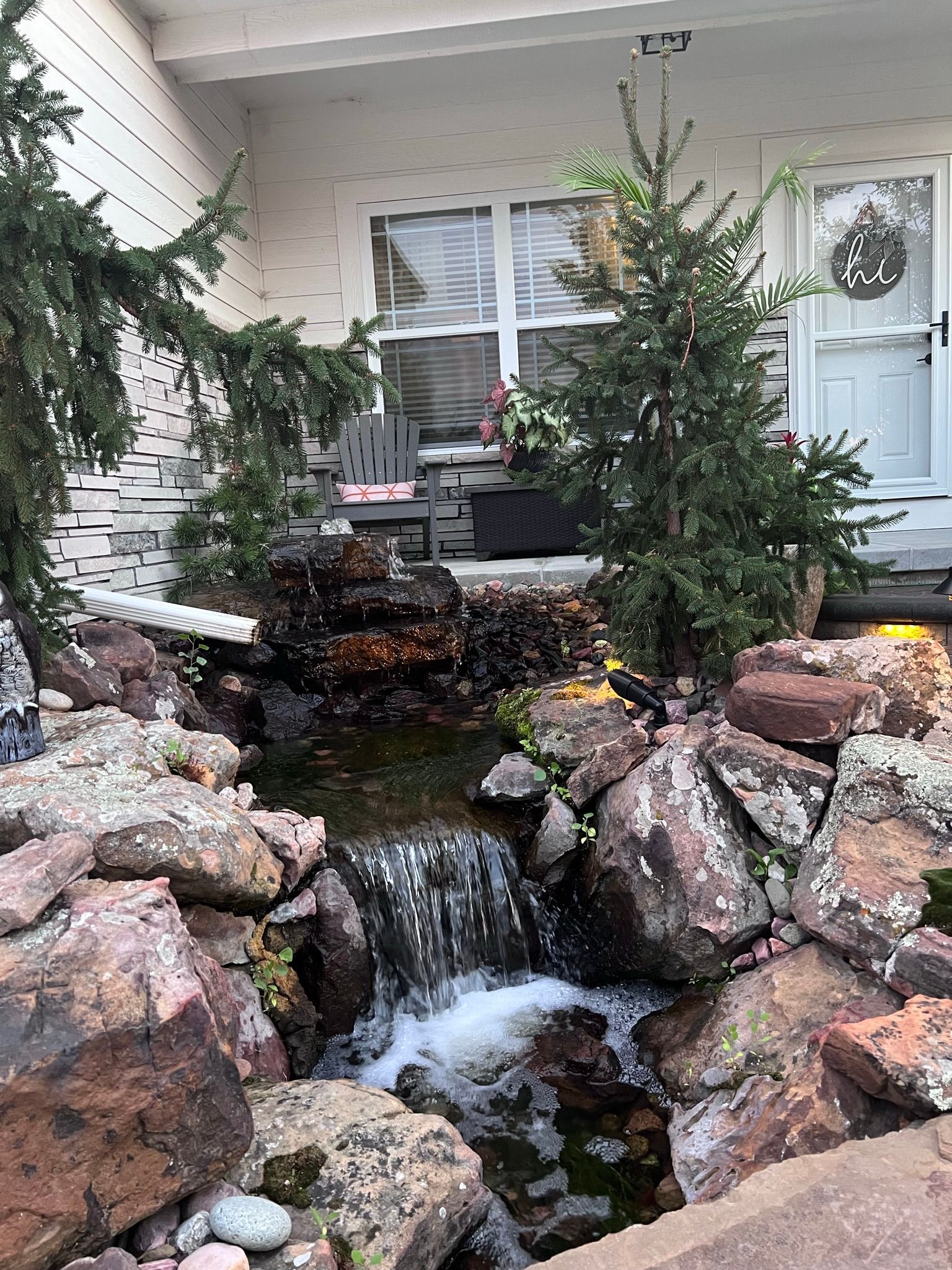 A small waterfall flows into a pond surrounded by large rocks, with a house in the background.
