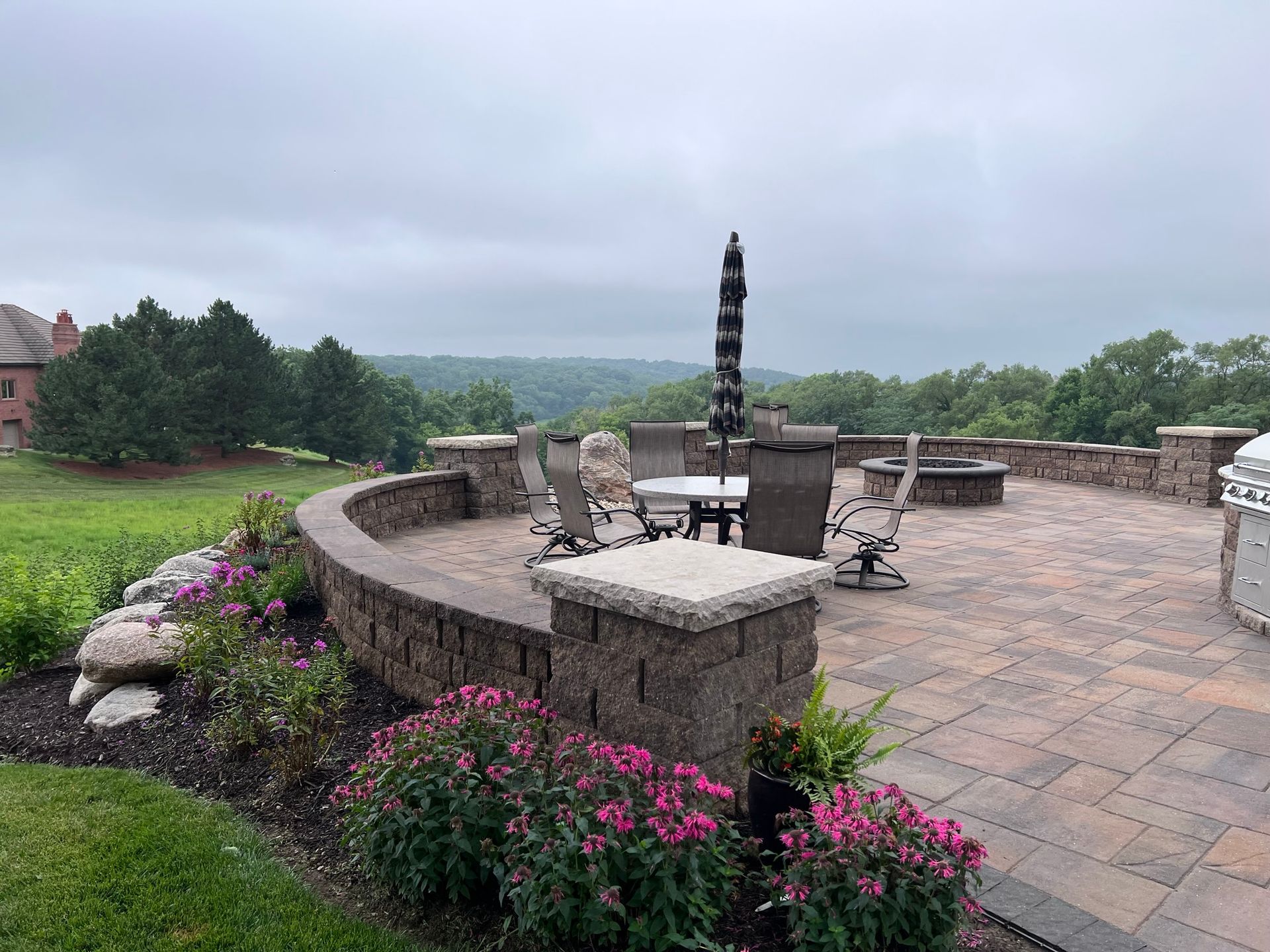 Patio with stone pavers, seating, fire pit, and umbrella, overlooking a wooded landscape.