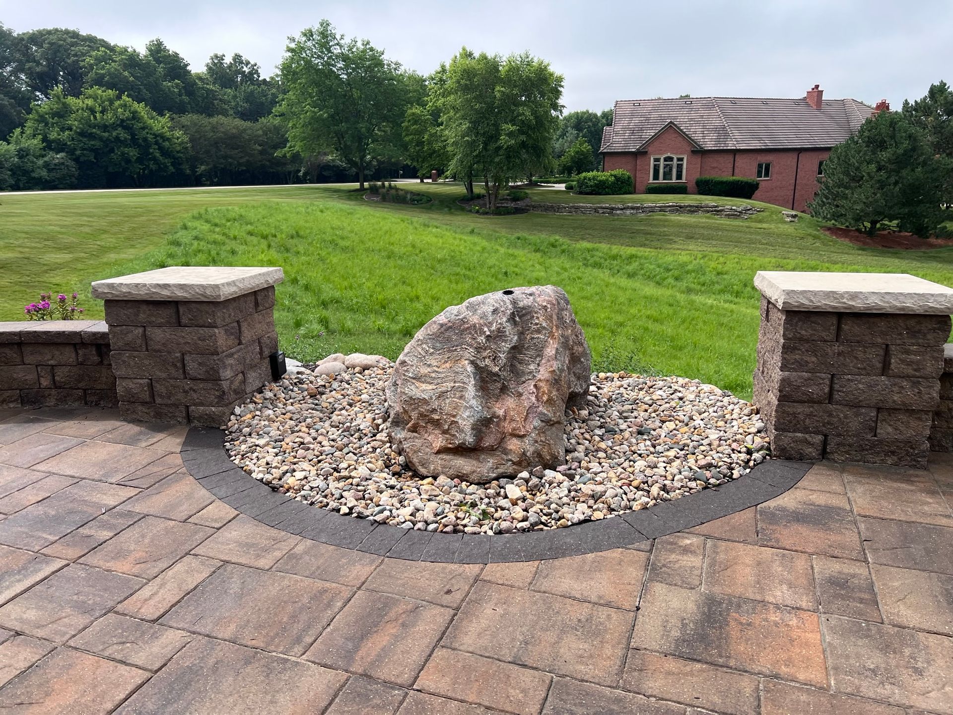A large boulder in a circular gravel bed on a paved patio, with two brick columns and a green, grassy landscape.